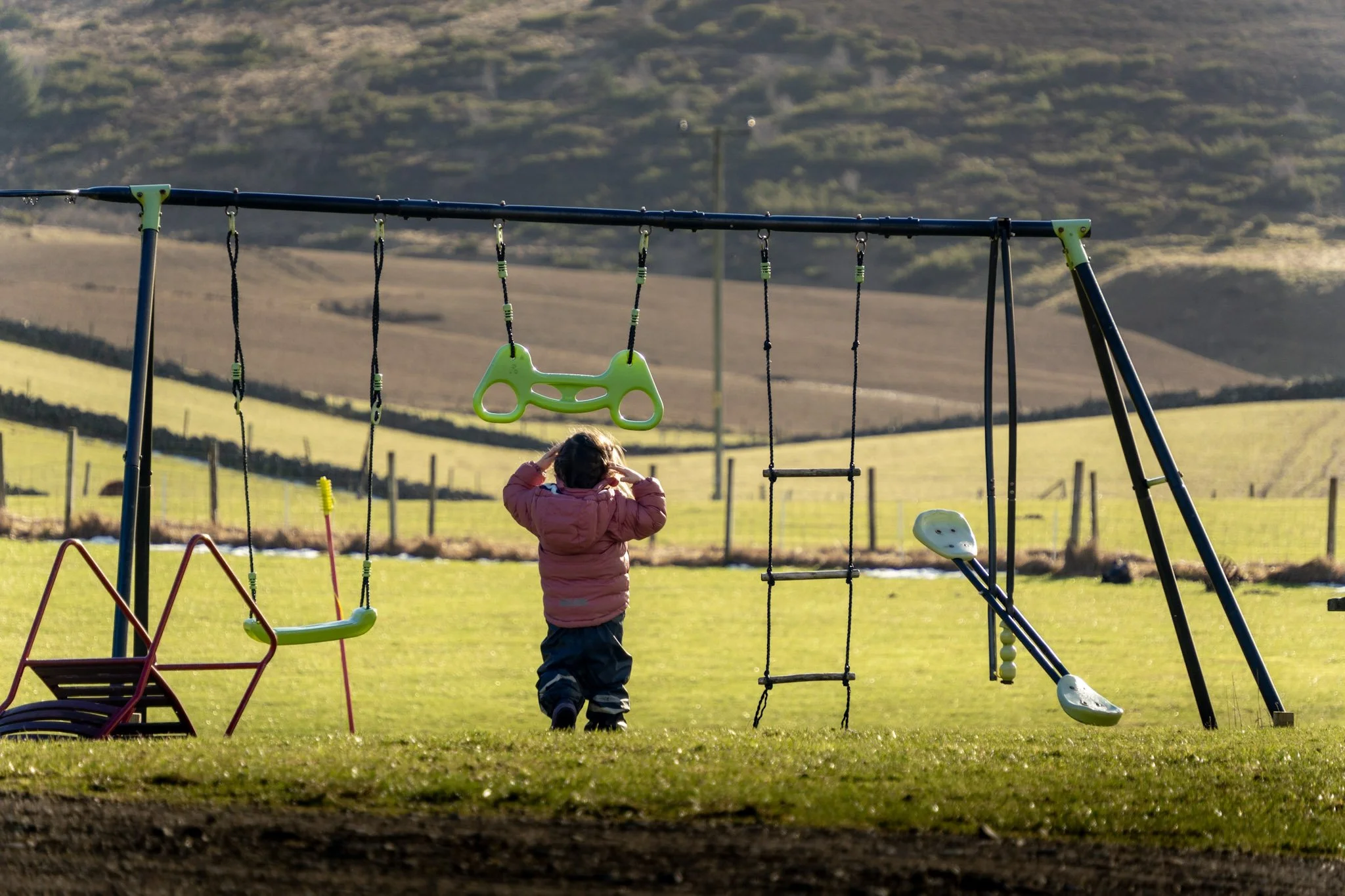 A young child in a pink jacket playing on swing sets in a grassy park area with rolling hills and fences in the background.