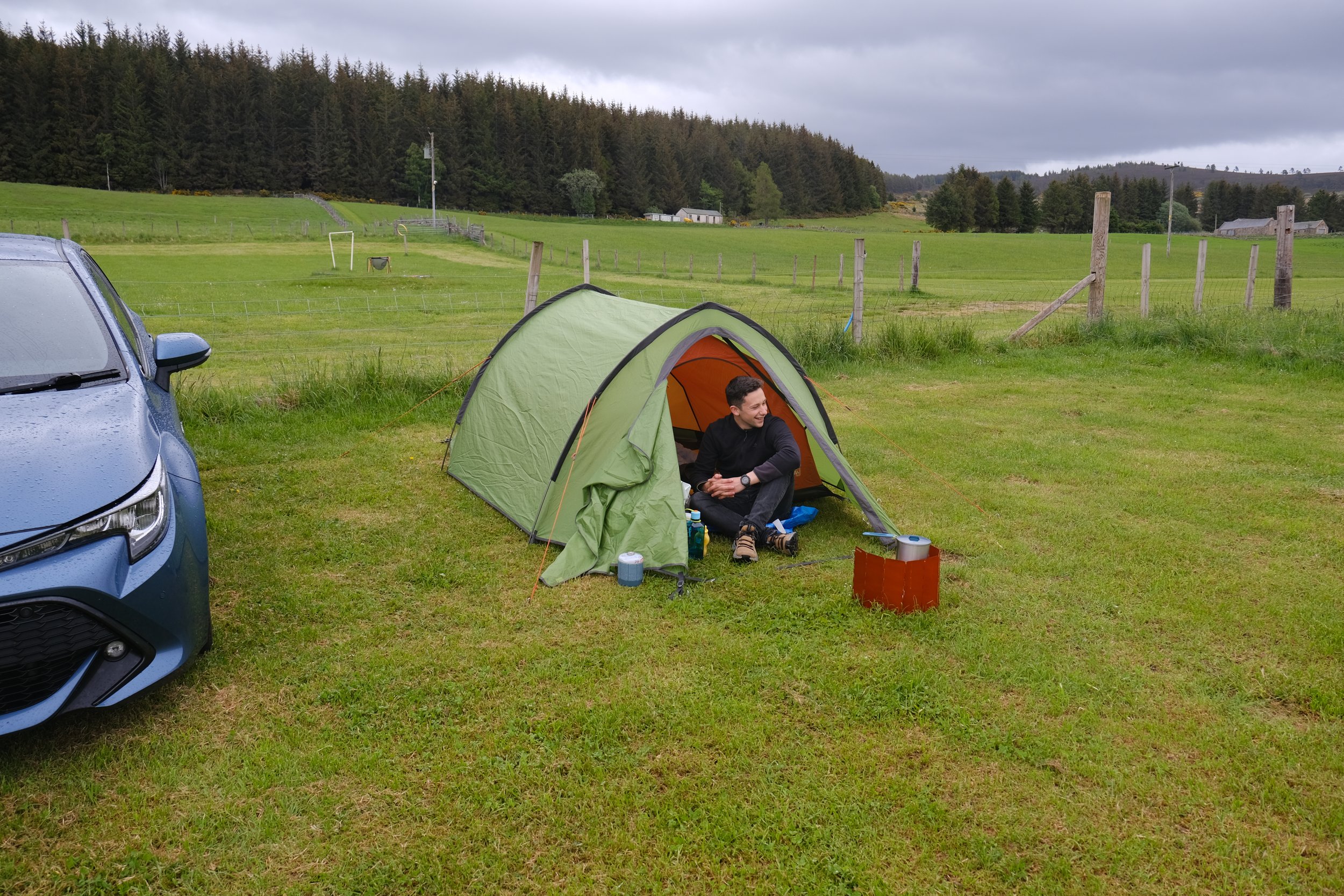A person sitting inside a green camping tent on a grassy field near a blue car, with a background of rolling green fields, trees, and a cloudy sky.