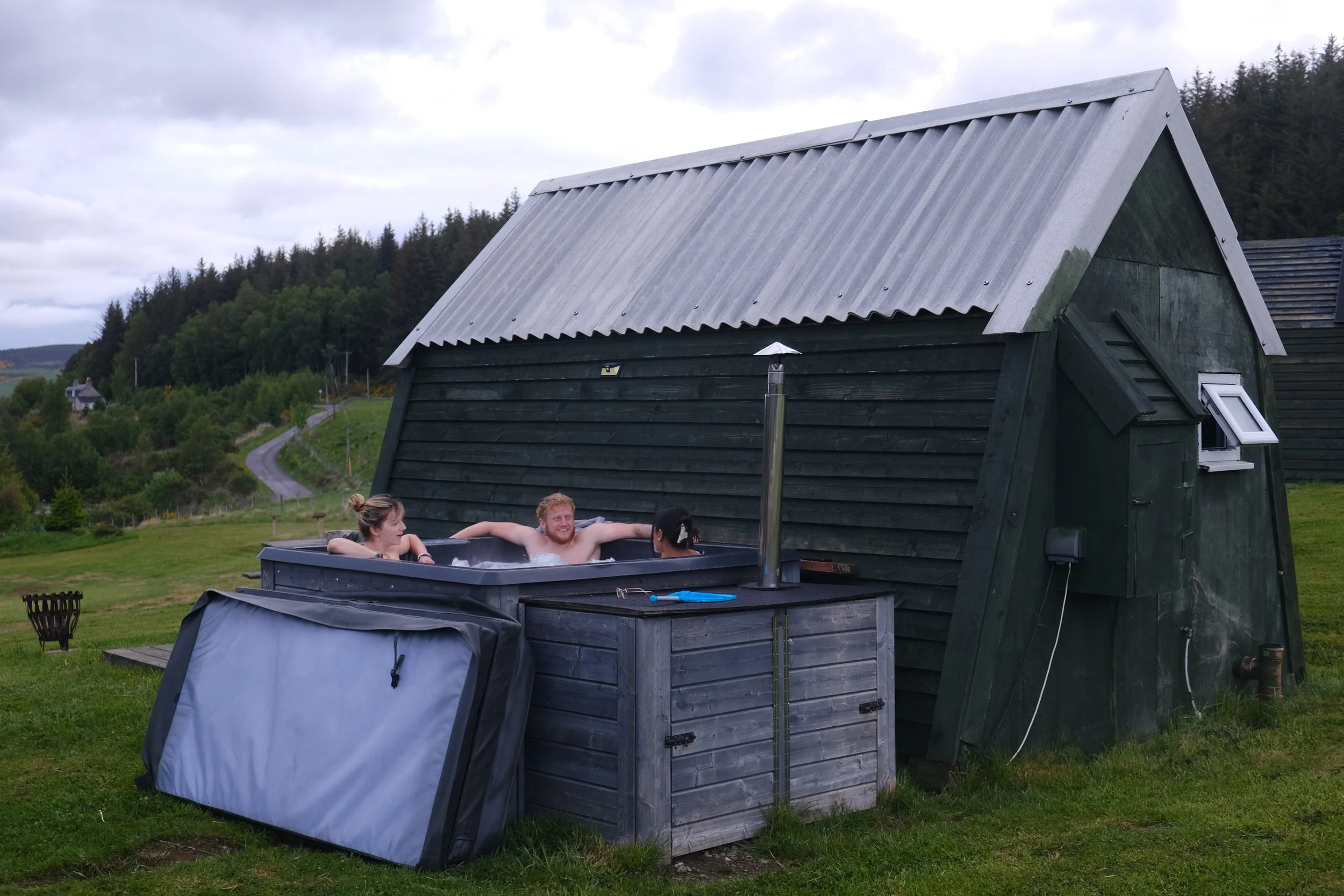 People relaxing in an outdoor hot tub in front of a dark green wooden cabin with a metal roof, with scenic hills and trees in the background.