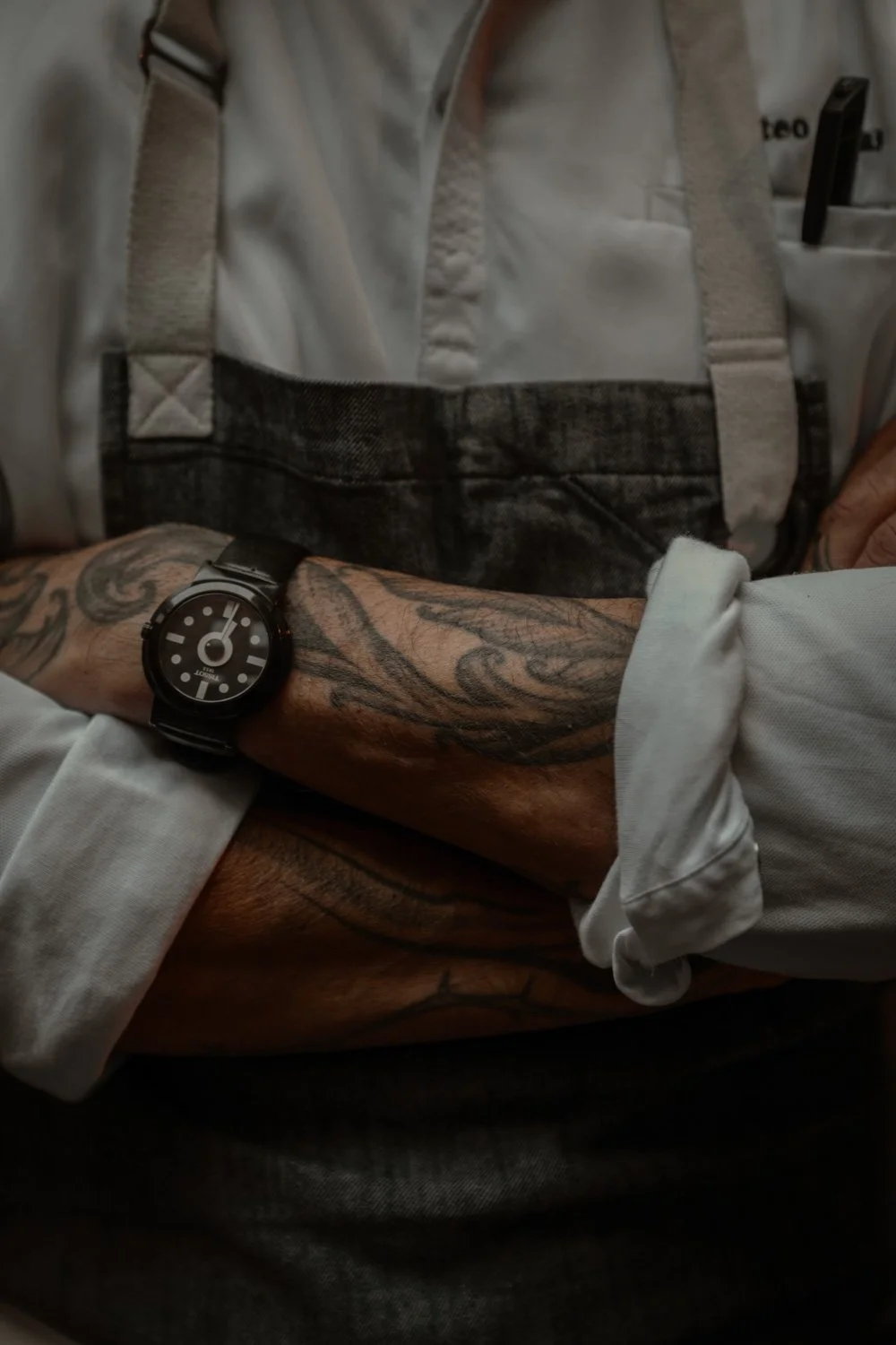 Nahaufnahme eines tätowierten Arms mit schwarzer Armbanduhr, getragen von einer Person in Berufskleidung, möglicherweise in einem Pflege- oder Serviceberuf.