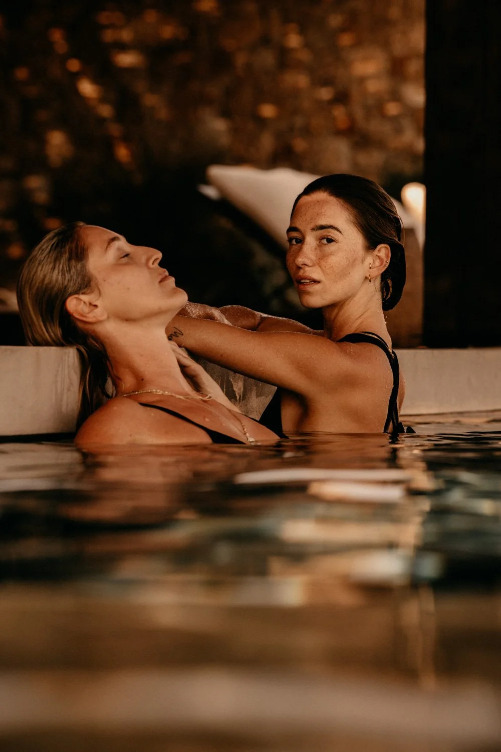 Zwei Frauen in einem warm beleuchteten Pool, eine streicht der anderen sanft über den Hals, im Hintergrund ist ein dunkler, unscharfer Raum sichtbar.