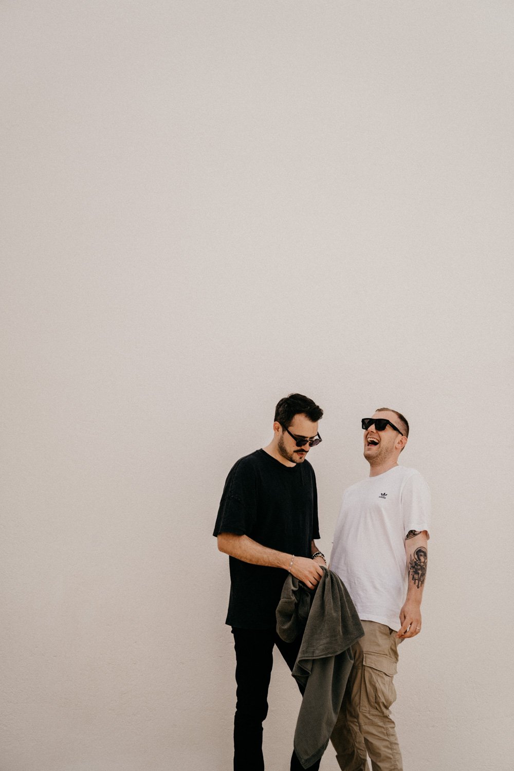 Zwei junge Männer lachen und haben Spaß, während sie vor einer weißen Wand stehen. Einer trägt eine schwarze T-Shirt und Sonnenbrille, der andere eine weiße T-Shirt, Sonnenbrille und hat ein Tattoo am Arm.