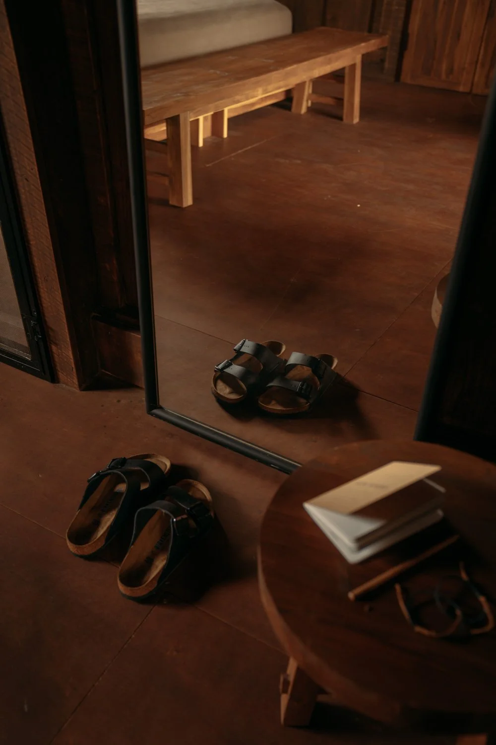 Zwei Paar schwarze Sandalen auf Holzboden vor einem Spiegel, mit einem runden Holztisch und einem Stapel Notizbücher.