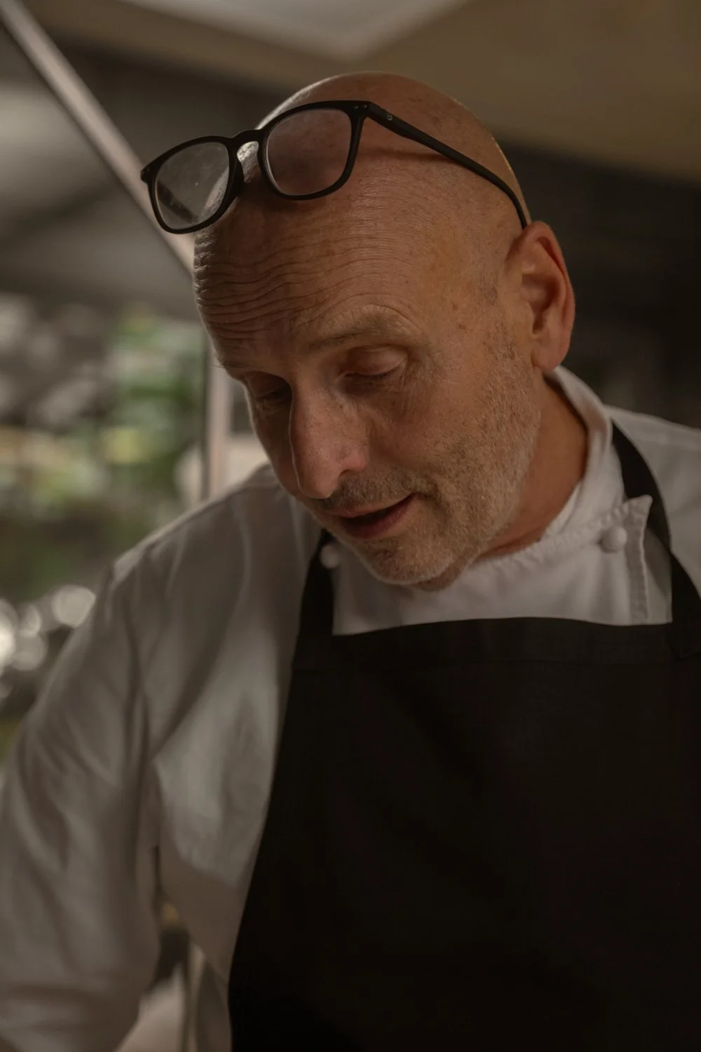 Ein älterer, braunhaariger Koch trägt eine weiße Kochjacke und eine schwarze Schürze. Er hat eine Brille auf dem Kopf, die er scheinbar gerade abgesetzt hat, und scheint in einem Restaurant oder Küchenbereich zu arbeiten.