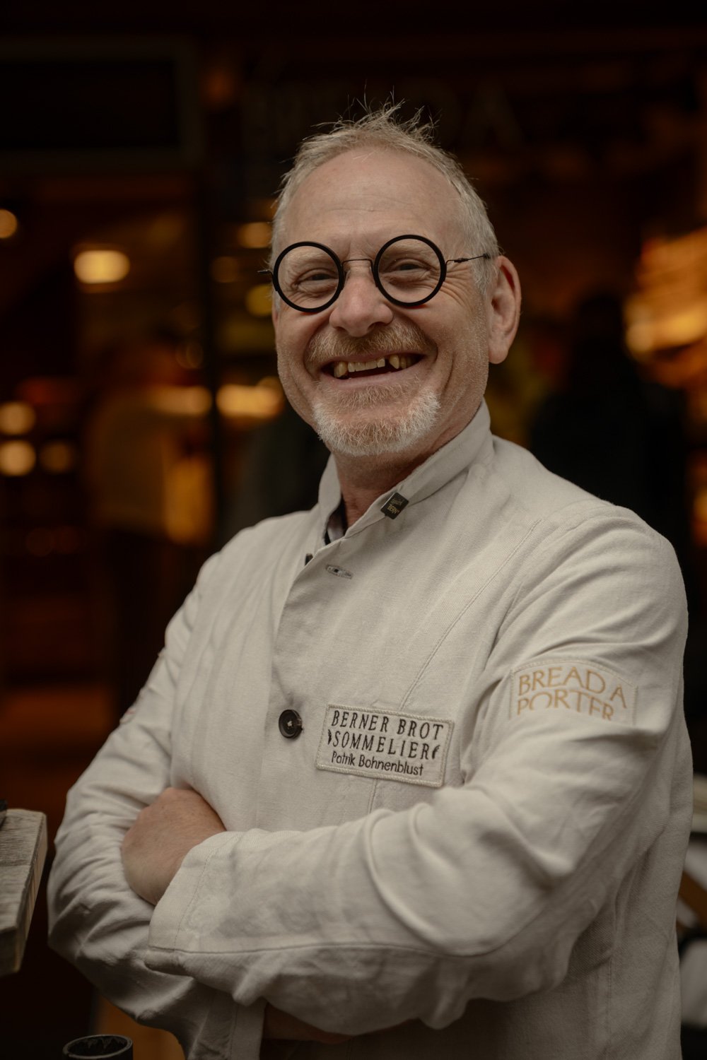 Ein lachender älterer Mann mit Brille und grauem Bart trägt eine weiße Kochjacke mit Namensschild, im Hintergrund ein Restaurant.