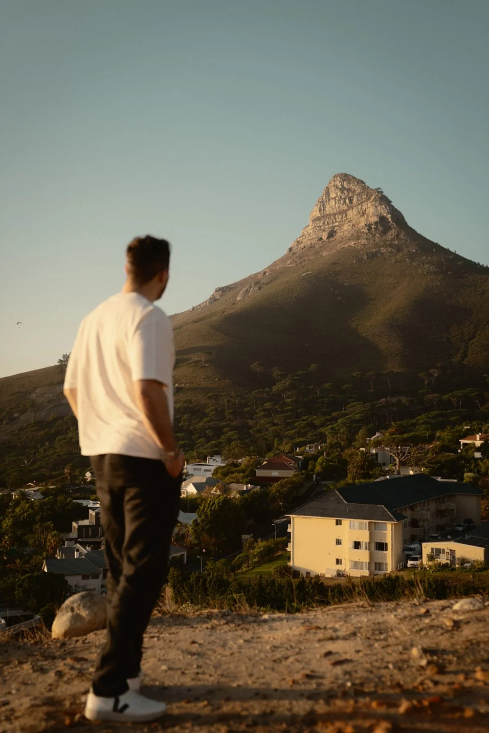 Ein junger Mann steht auf einem Hügel und blickt auf eine Stadt mit Häusern und Bäumen, im Hintergrund ein Berg bei Sonnenuntergang.
