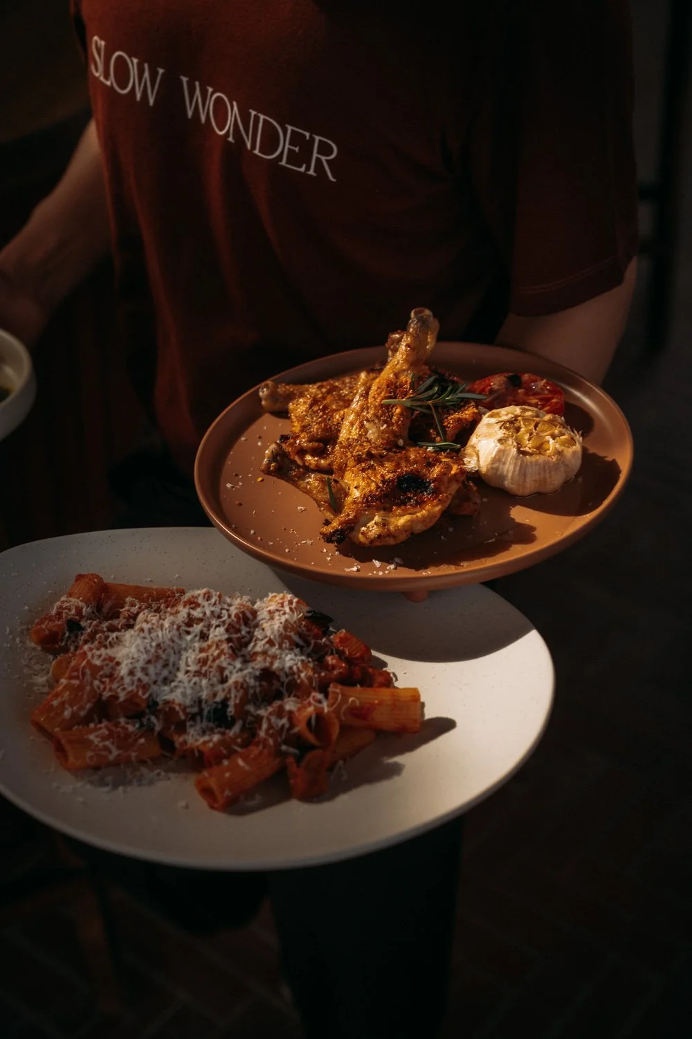 Eine Person hält eine braune Platte mit gebratenen Hähnchenteilen, Knoblauch und Tomaten. Vor der Person liegt eine weiße Platte mit Pasta und Tomatensauce, bestreut mit Parmesan.