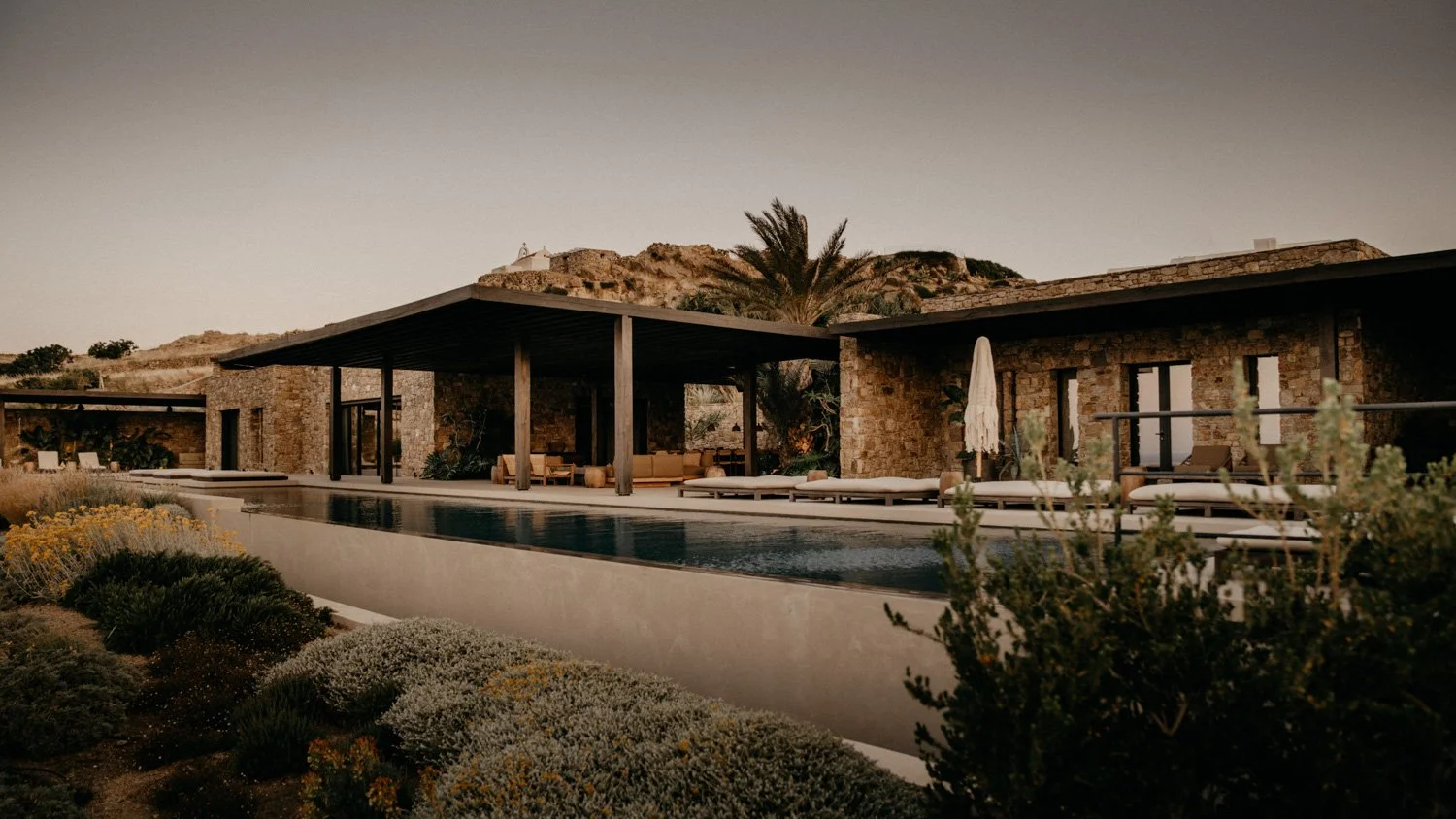 Modernes Haus mit Steinwänden, Pool im Vordergrund, Sonnenschirm und Liegestühle am Pool, bewaldete Hügel im Hintergrund, bei Sonnenuntergang.