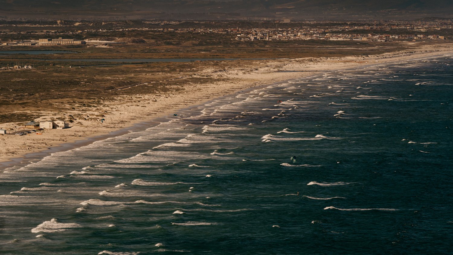 Küstenabschnitt mit Sandstrand und Wellen, im Hintergrund Stadtlandschaft