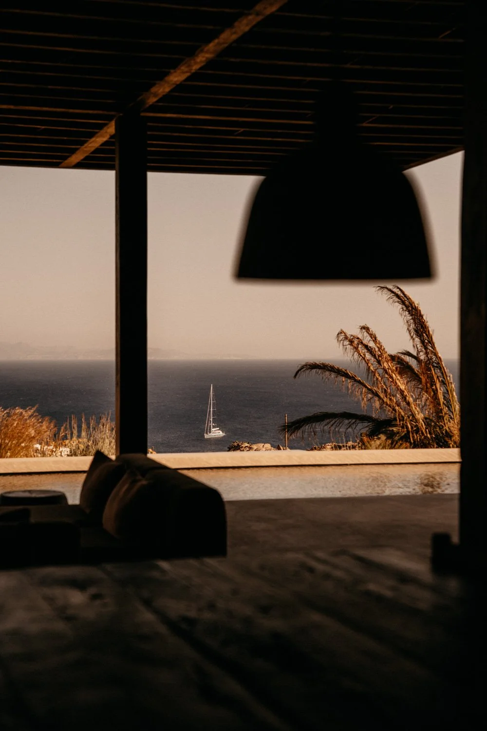 Blick auf das Meer mit Segelboot durch eine offene Terrasse mit hängender Lampe und Pflanzen im Vordergrund, Sonnenuntergangsstimmung.