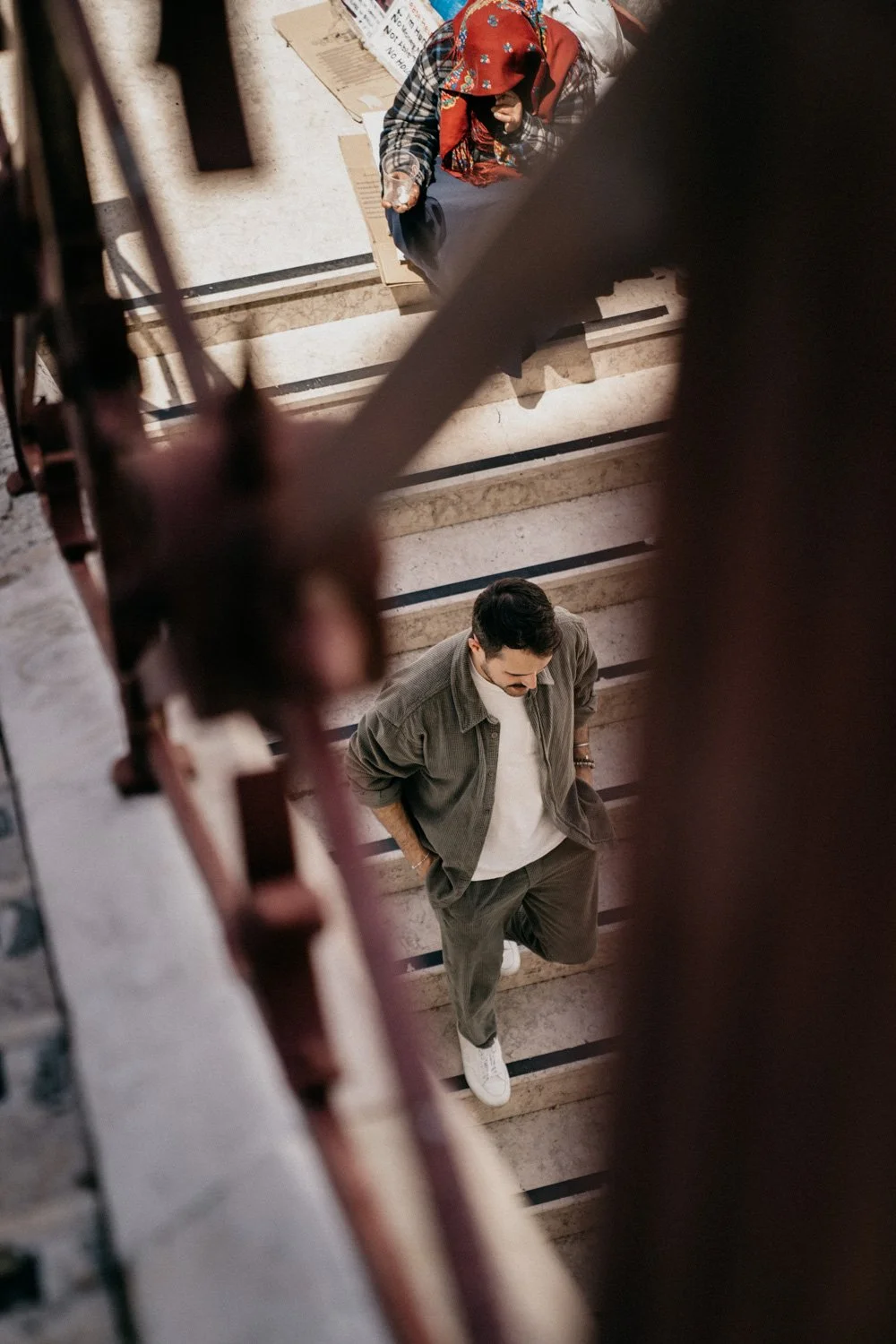 Ein Mann in einem grauen Anzug und weißer Sneakers, der eine Treppe hinuntergeht, betrachtet nach oben, während eine Frau mit einem roten Schal und einer Jacke auf dem Boden sitzt.