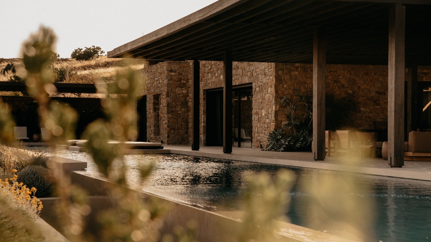 Moderne Villa mit Steinfassade, Holzstützen, Pool und umgebender Natur bei Sonnenuntergang.