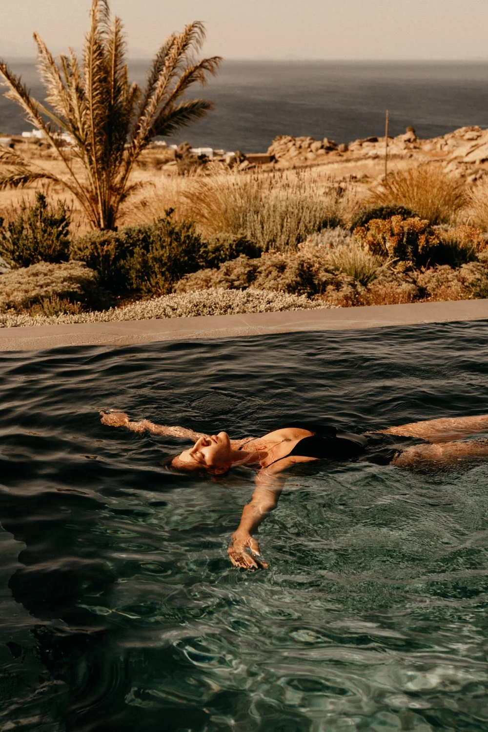 Frau entspannt im Pool liegend mit Blick auf eine trockene, mediterrane Landschaft mit Palmen und Blick aufs Meer im Hintergrund.