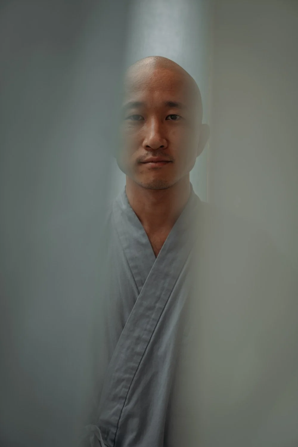 Mann in grauem Kimono, schaut durch eine kleine Öffnung, vermutlich durch eine Tür.