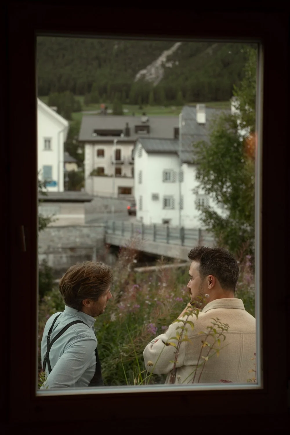 Zwei Männer, die durch ein Fenster von innen nach draußen schauen, während sie sich in einer Gartenumgebung unterhalten. Im Hintergrund sind weiße Häuser, eine Brücke und ein bewaldeter Hügel sichtbar.