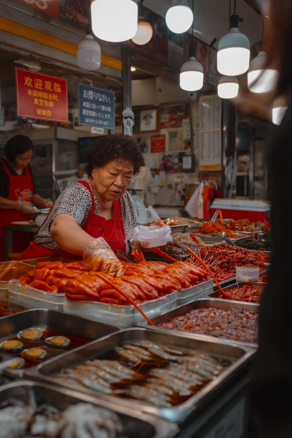 Eine ältere Frau verkauft koreanisches koreanisches Street Food, darunter marinierte Meeresfrüchte und scharf gewürzte Saucen in einem Markt.