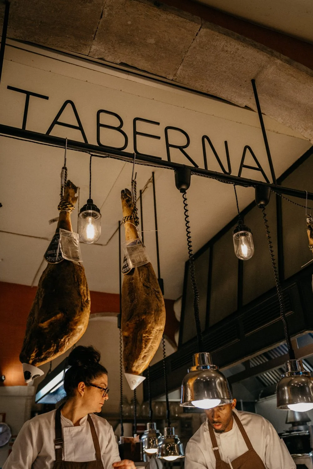 Barg, auf dem die aufgehängte Schilder mit der Aufschrift 'TABERNA' zu sehen sind, mit zwei Köchen, die in der Küche arbeiten, und an der Decke hängen Schinken, Lampen und Kabel.