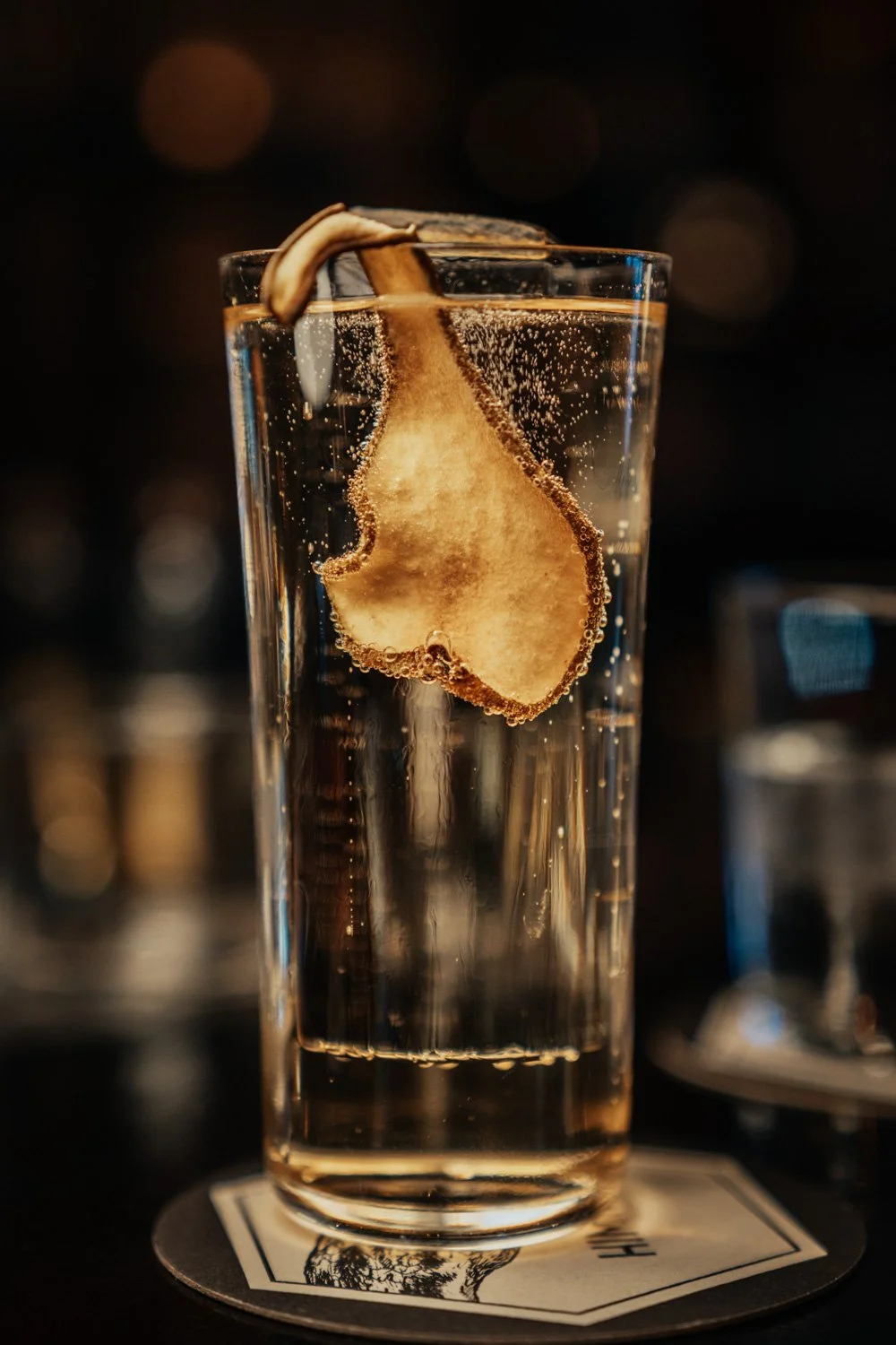 Ein hohes Glas mit Wasser und einer Zitronenscheibe, mit Blasen, auf einem Untersetzer im Dunkeln.