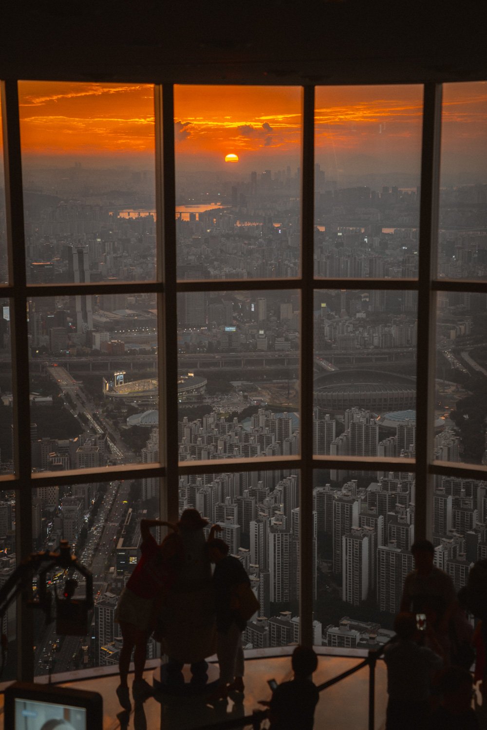 A sunset over a cityscape viewed through a large window with gridpanes, with silhouettes of people in the foreground.