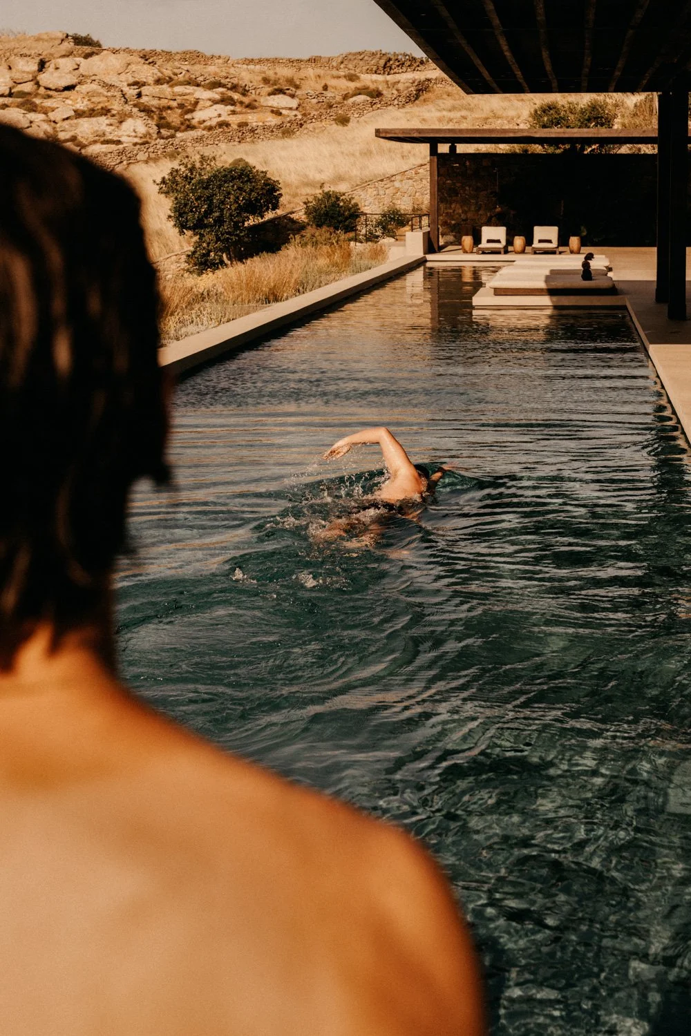 Person schwimmt im Pool während eine Person im Vordergrund zuschaut, in einer modernen, stilvollen Pool-Umgebung mit Sitzgelegenheiten und einem ländlichen Bergblick im Hintergrund.