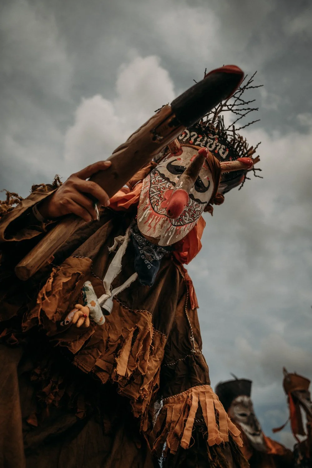 Eine Person trägt eine große, kunstvolle Maske und ein Kostüm aus Holz und Stoff, während sie einen Holzkeulen hält. Der Himmel im Hintergrund ist bewölkt.