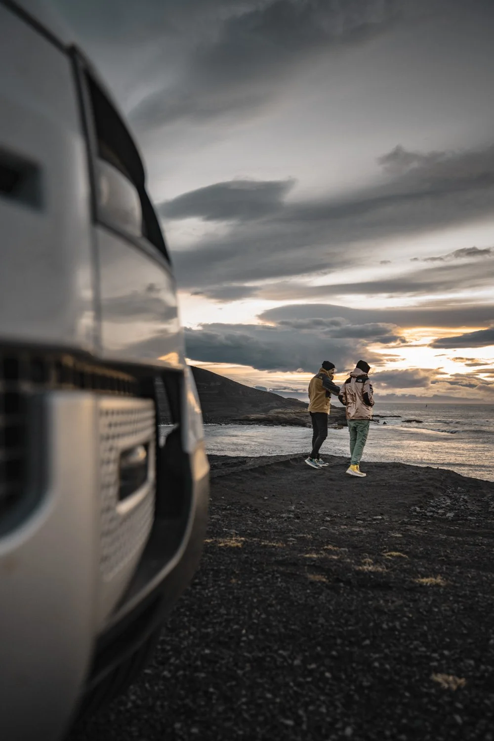 Zwei Personen am Strand bei Sonnenuntergang, im Hintergrund das Meer und bewölkter Himmel, links ein Teil eines weißen Autos.
