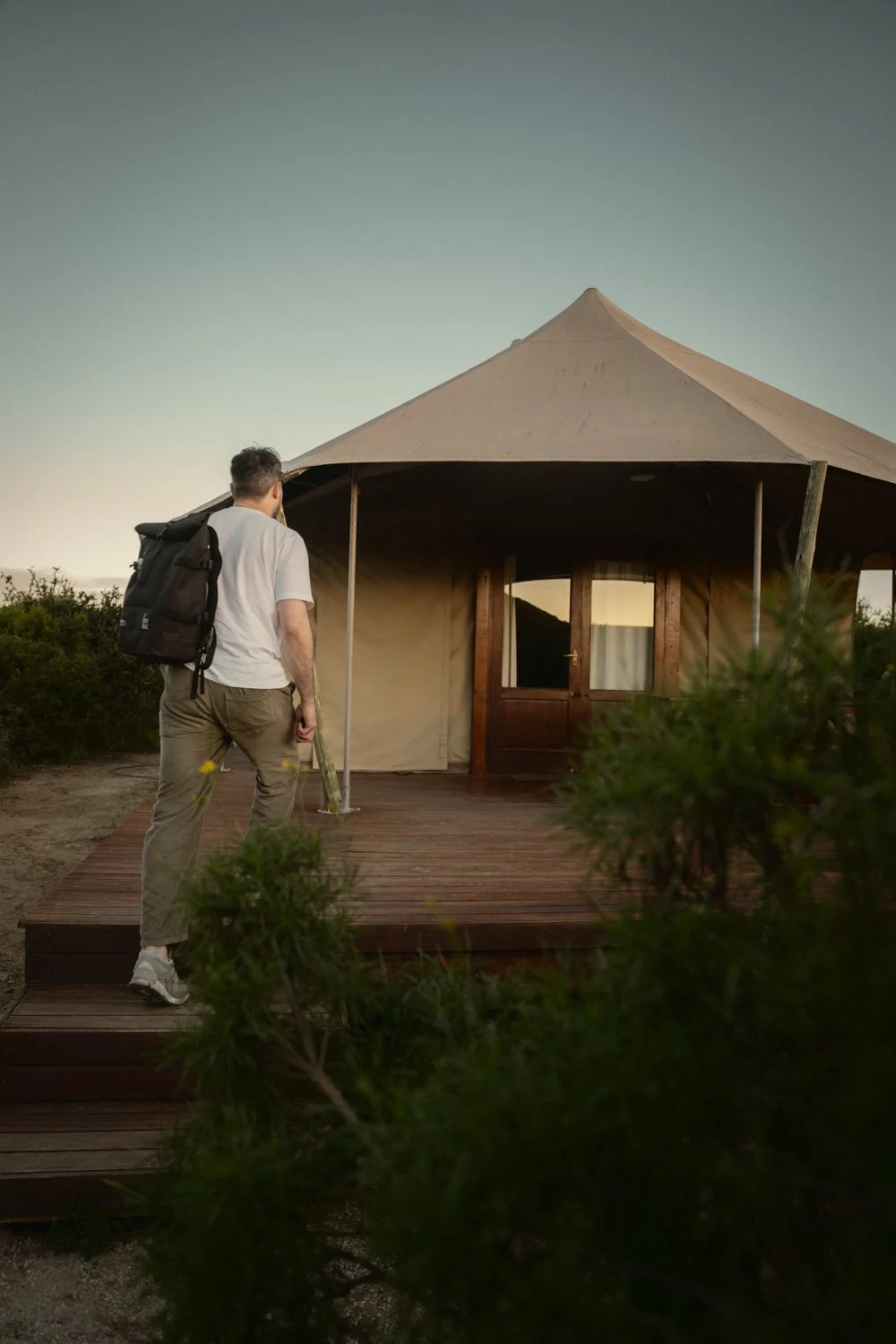 Ein Mann mit Rucksack steht vor einem runden Zelt auf einer Holzplattform, umgeben von Natur, bei Sonnenuntergang.