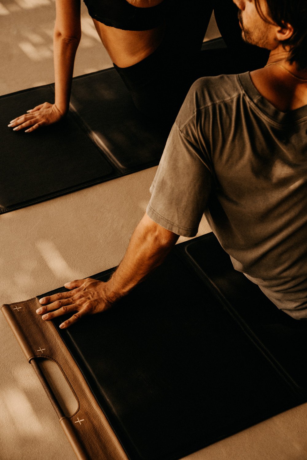 Zwei Menschen, die Yoga oder Pilates auf schwarzen Matten machen, eine Person in schwarzen Kleidung und die andere in grauem T-Shirt, in einem Raum mit gedämpftem Licht.