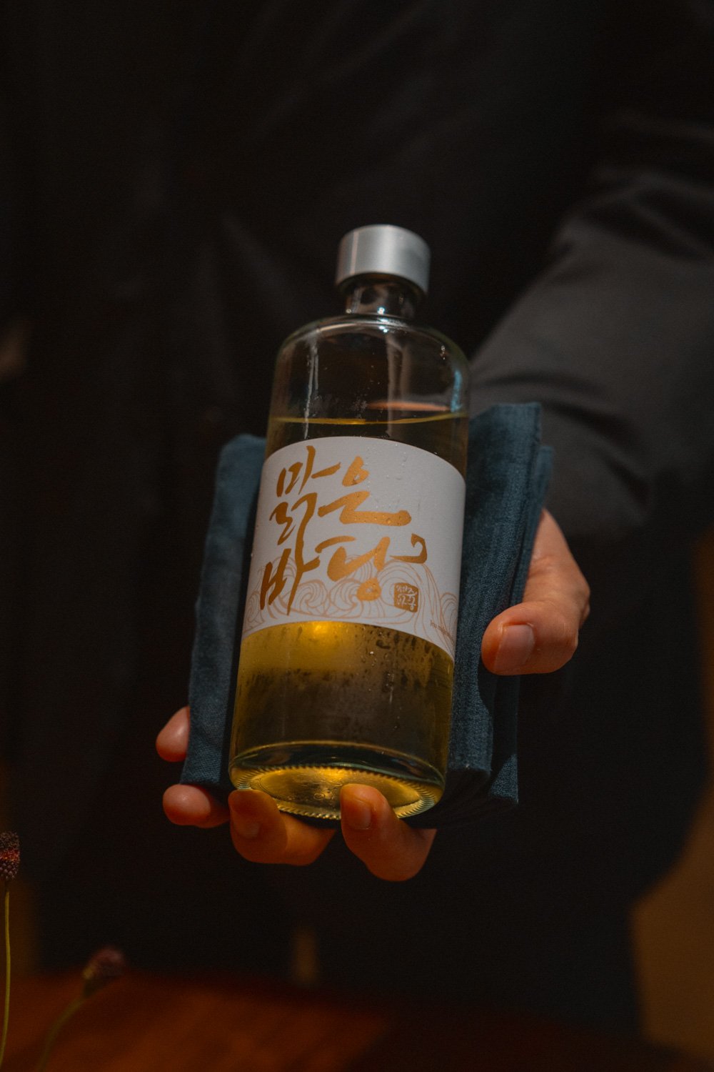 Hand hält eine Flasche mit hellem Getränk und asiatischem Schriftzug auf einem weißen Etikett, die in eine Hand mit dunklem Handtuch wickelt.