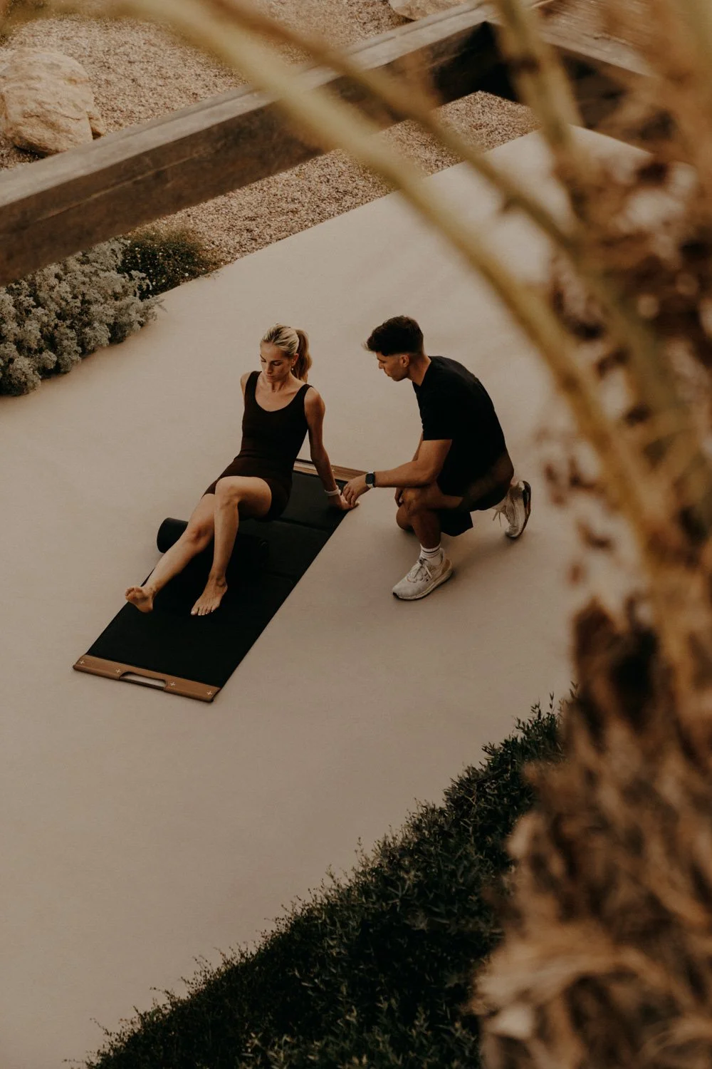 Ein Mann hilft einer Frau, die auf einer Yogamatte sitzt, draußen bei Sonnenuntergang.