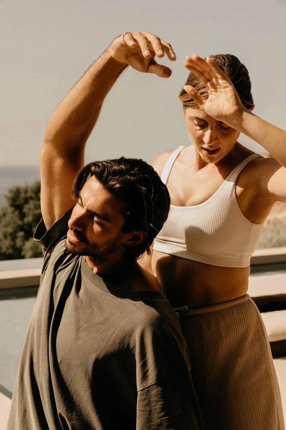 Zwei Menschen beim Yoga auf einer Terrasse mit Meer im Hintergrund, Sonnenlicht, entspannt.