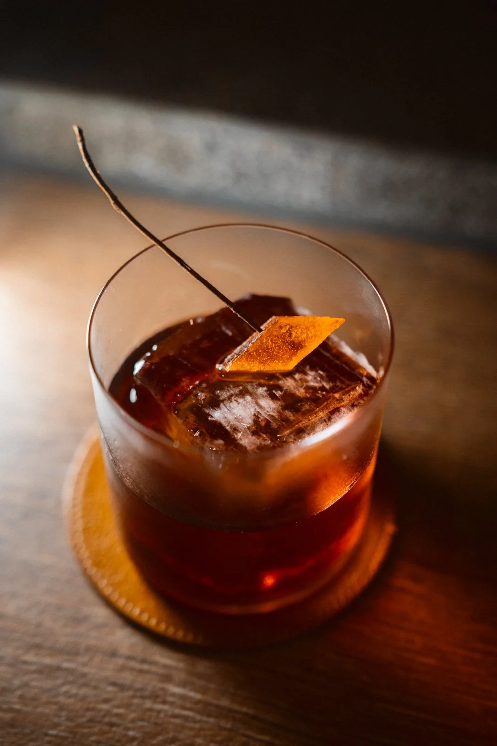 Ein Glas mit einem dunklen alkoholischen Getränk, Eiswürfeln und einer Orangenscheibe als Garnitur, auf einem Holzuntergrund.