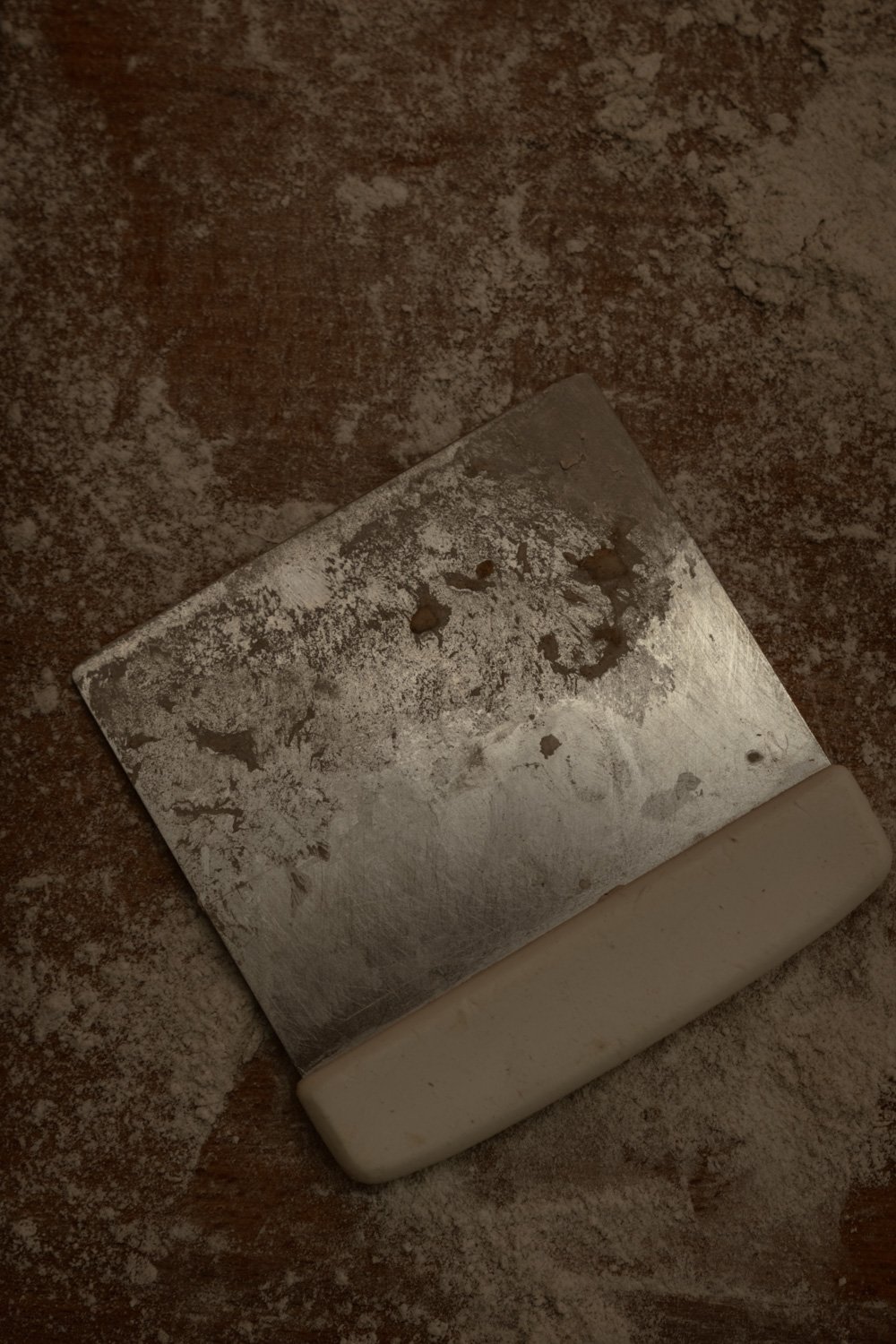 Ansicht eines Metallrakels auf einem groben, staubigen Untergrund, mit einem weißen Griff am unteren Rand.