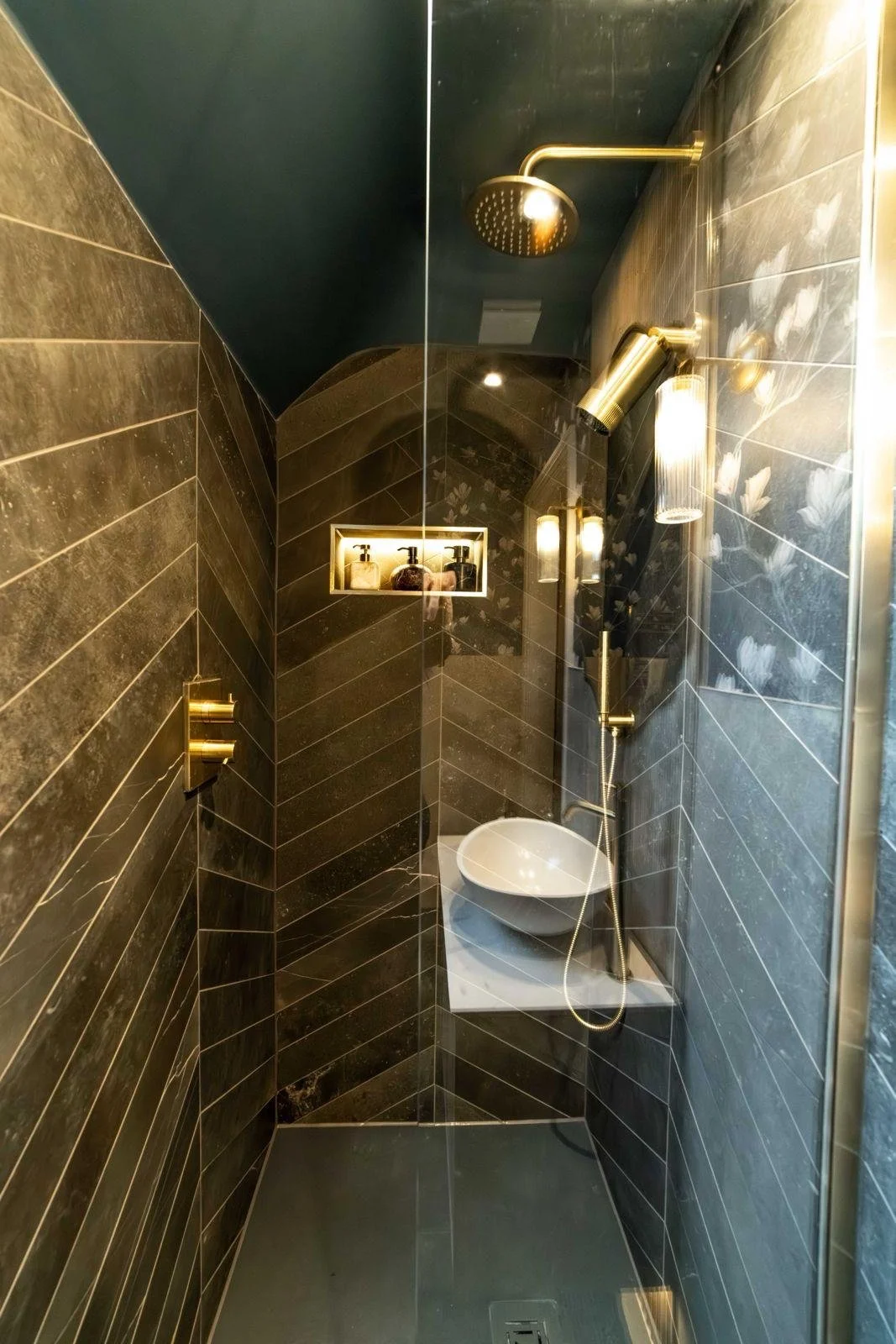 Small modern shower with black and gold accents, a rain shower head, a wall-mounted shower head, a built-in shelf with bath products, and a small white basin.