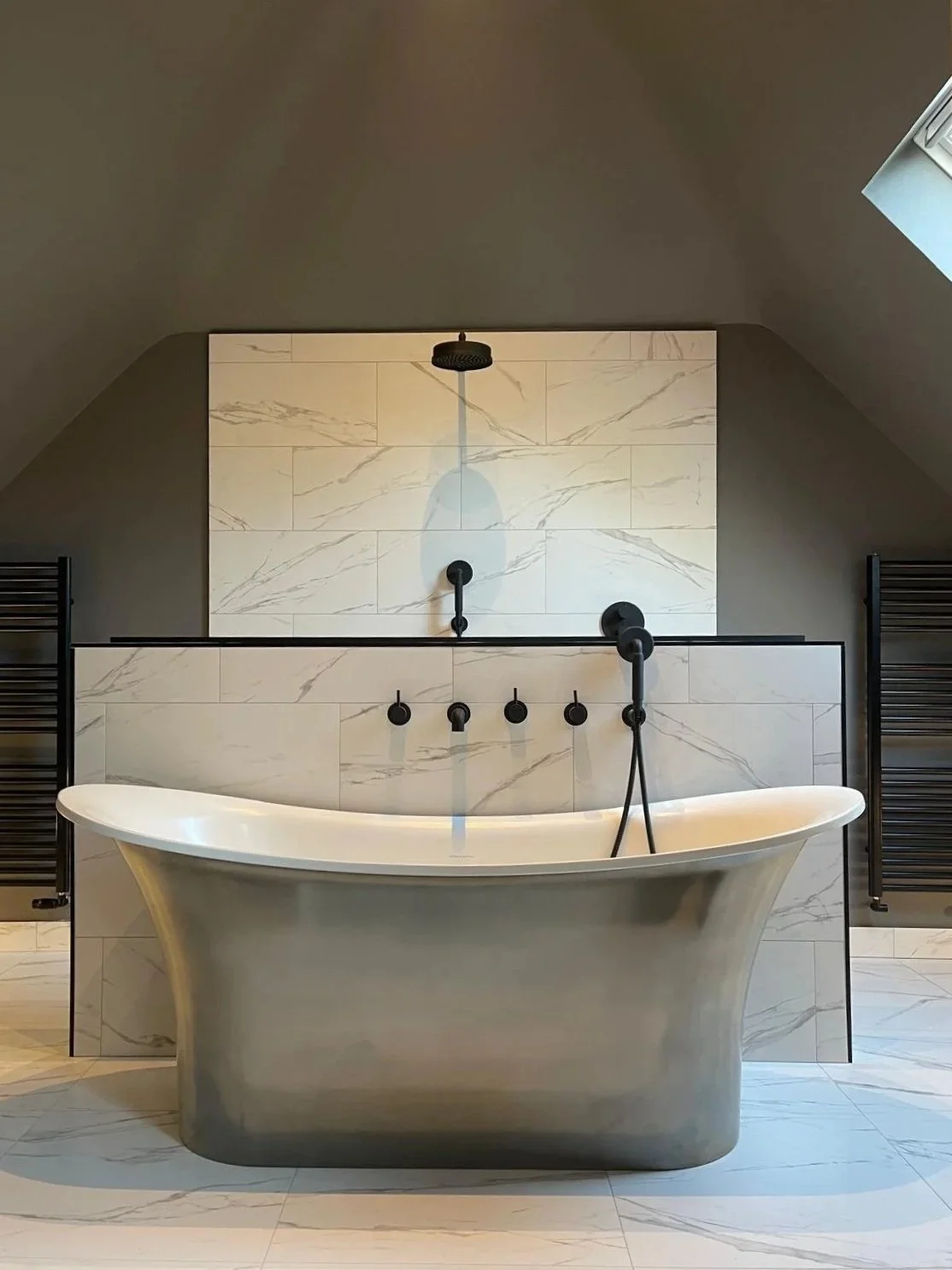 Modern bathroom with a freestanding bathtub, black fixtures, and marble wall tiles on an angled ceiling with a skylight.
