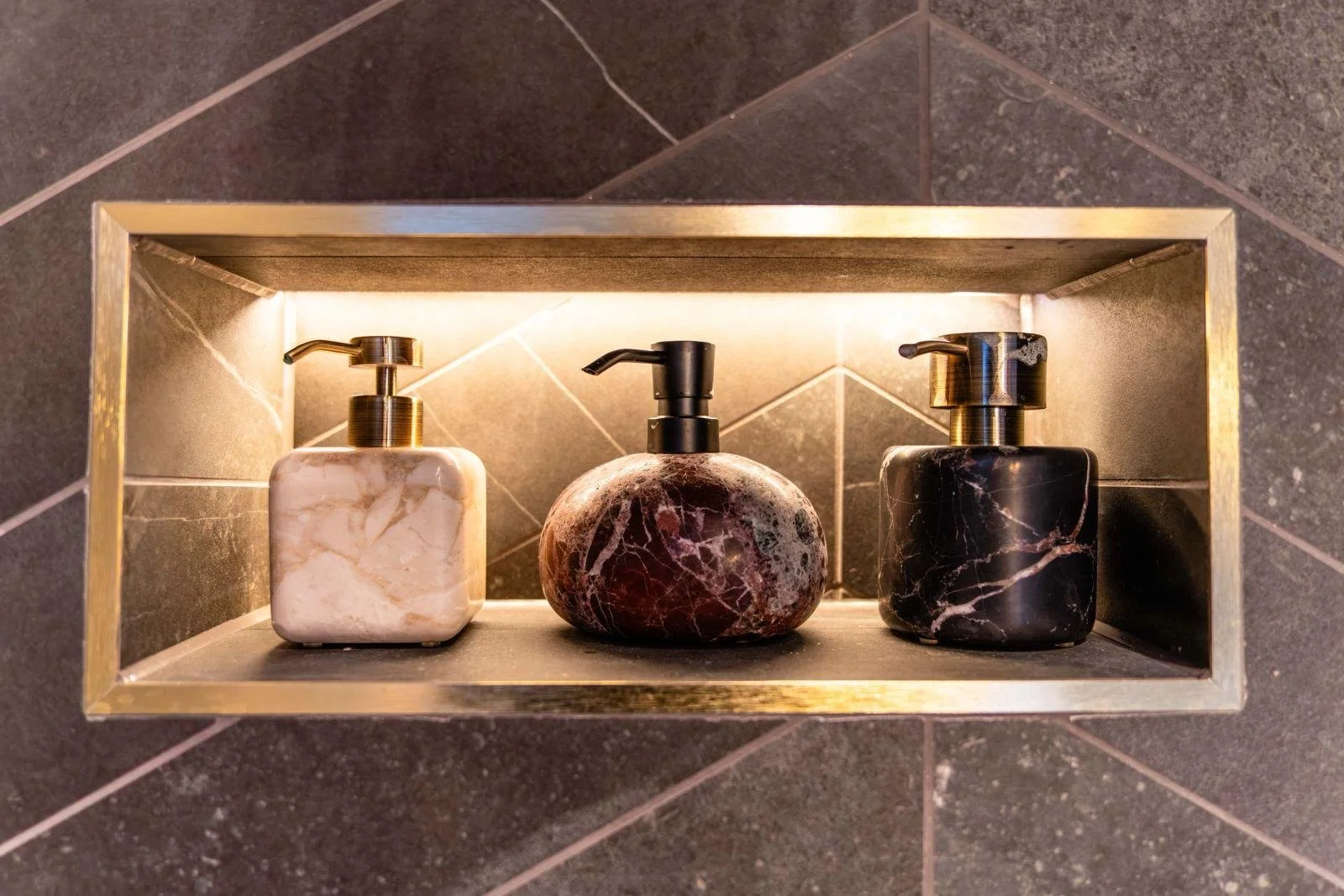Three marble soap dispensers on a shelf in a tiled shower or bathroom wall, with one white, one red, and one black marble finish.