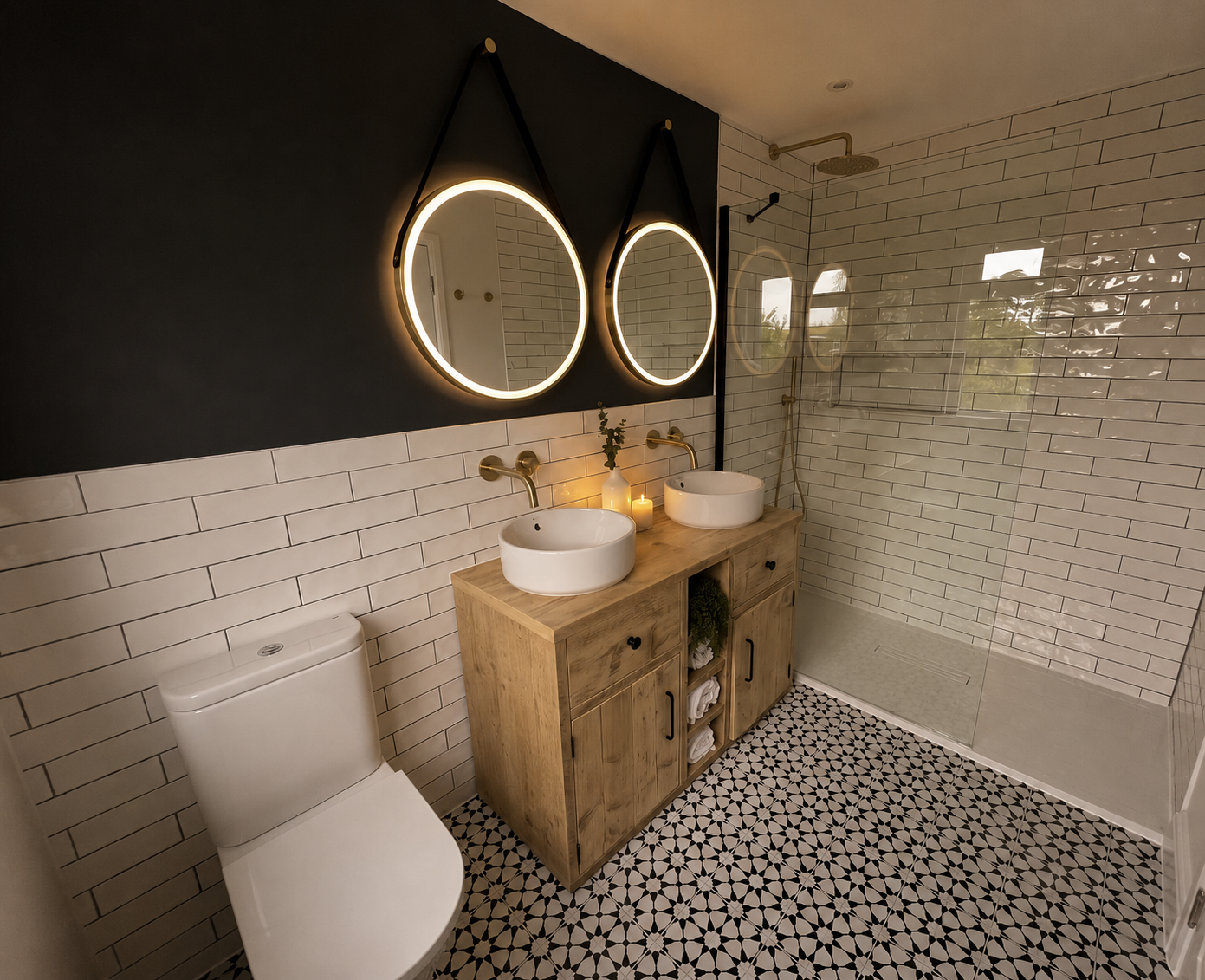Modern bathroom with two oval illuminated mirrors, double vessel sinks on a wooden vanity, black and white patterned tile floor, and a walk-in shower with white subway tiles and a glass partition.