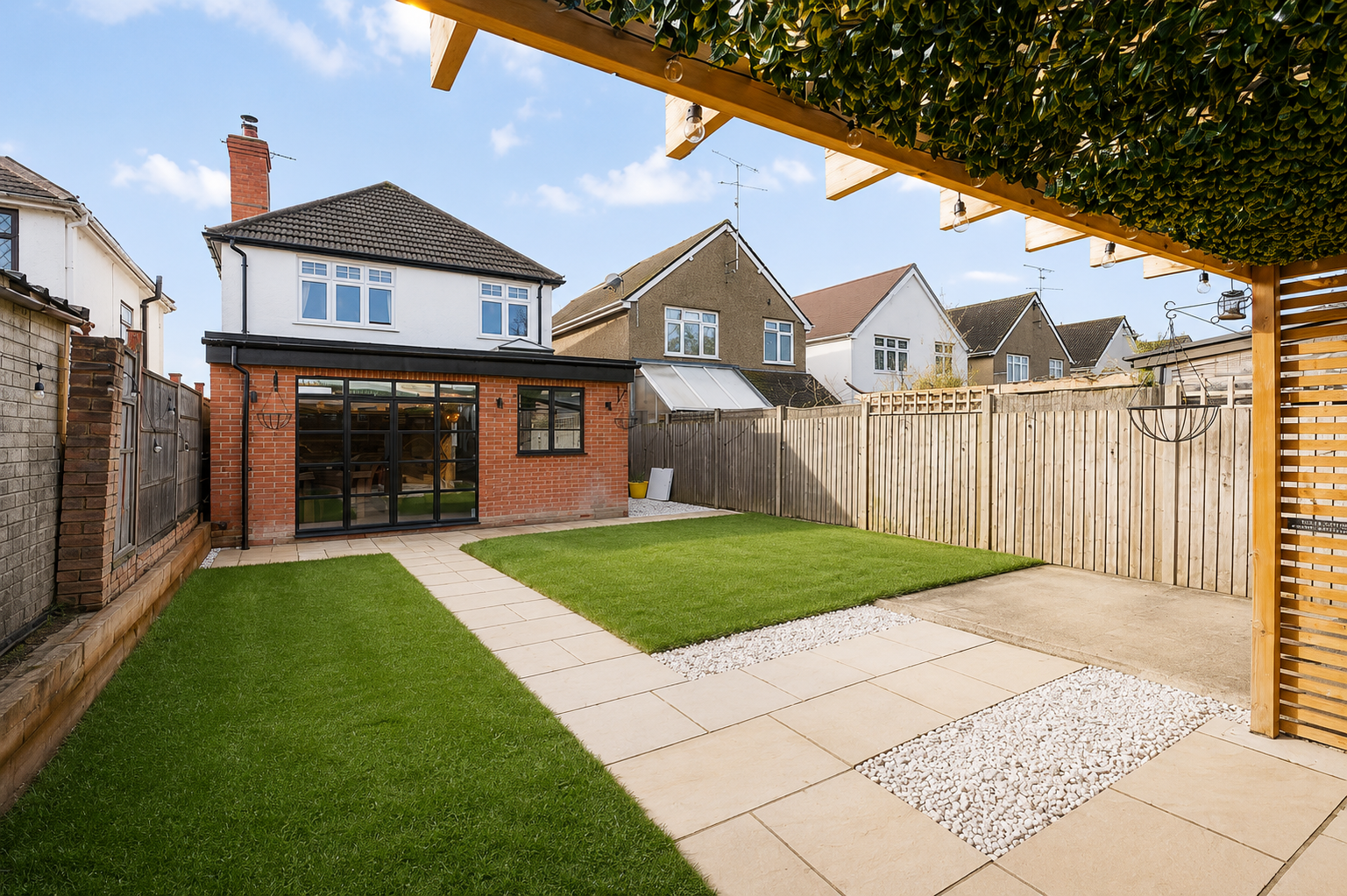 A backyard with a grassy lawn, paved walkway, and a wooden fence, with houses in the background under a blue sky.