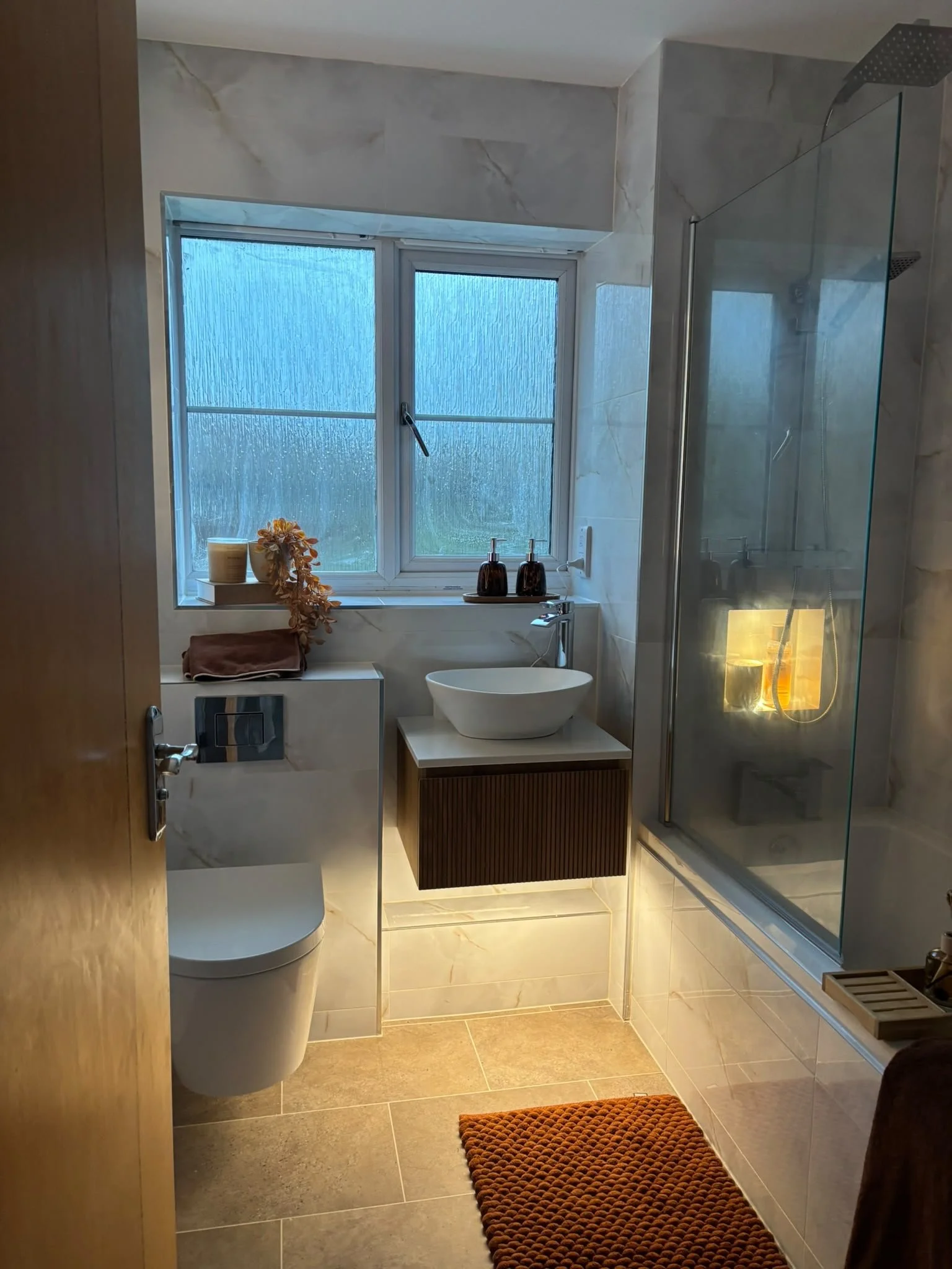 A modern bathroom with a window, a white toilet, a small wooden cabinet, a vessel sink, a shower with glass partition, and a rust-colored bath mat on beige tiled floor.