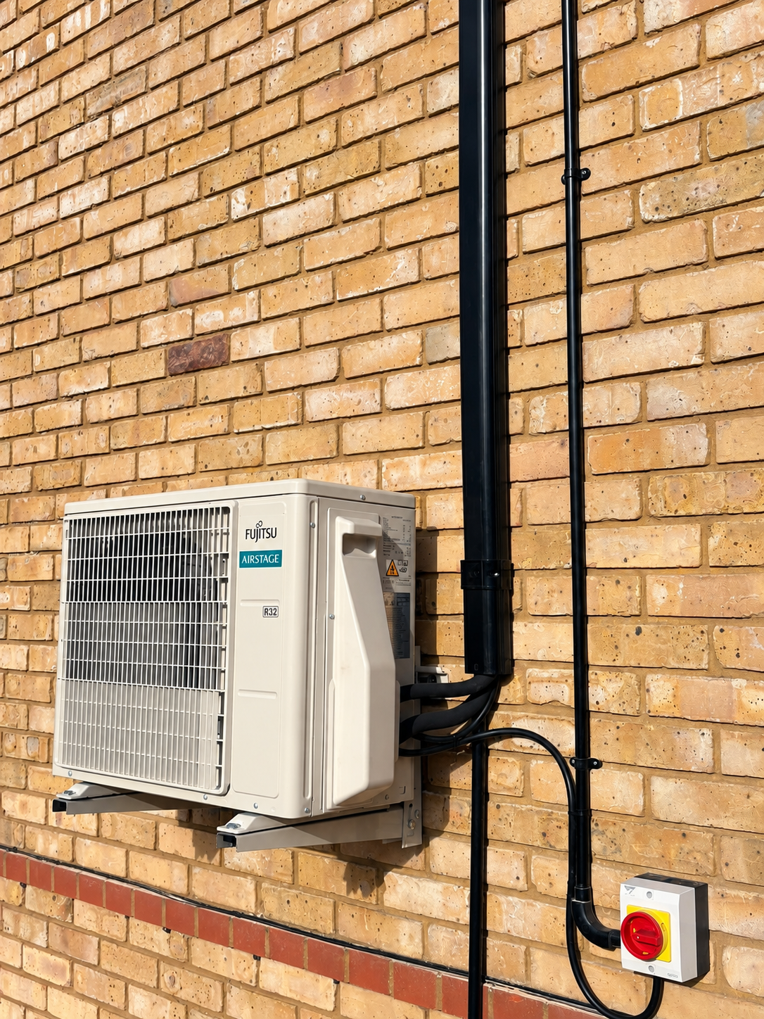 Outdoor air conditioning unit mounted on brick wall with electrical wiring and switch.