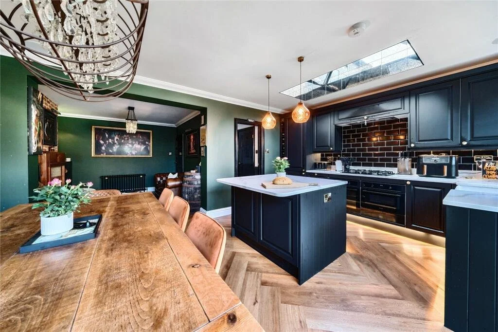 Open-concept kitchen and dining area with dark blue cabinets, white countertops, black backsplash, and wooden accents. The kitchen has a central island with pendant lights, and the dining table is large with a potted plant centerpiece.