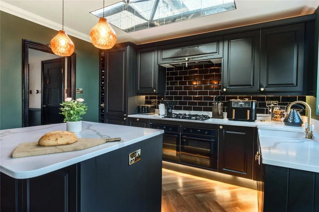 Modern kitchen with black cabinets, white countertops, black subway tile backsplash, kitchen appliances, and a center island with a loaf of bread and a potted plant.
