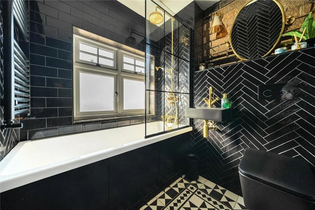 Modern bathroom featuring black tile walls with gold fixtures, a white bathtub, a small black sink with gold faucet, a round gold mirror, black and white patterned floor, frosted window, and gold accents.
