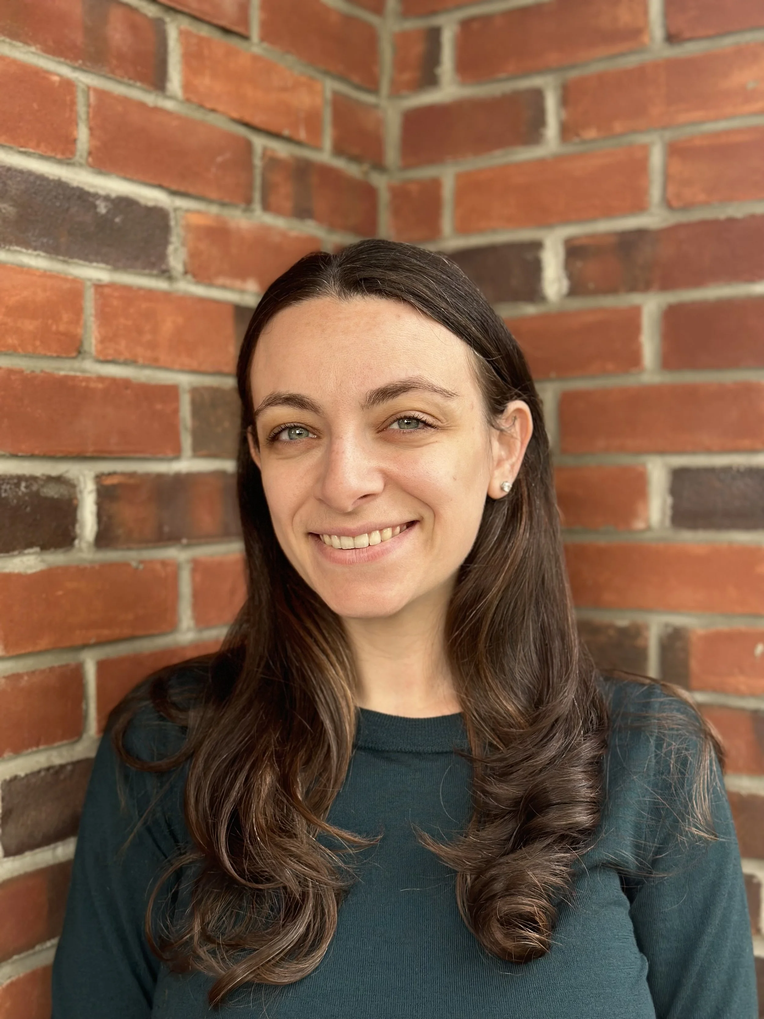A woman with dark brown hair smiling, standing in front of a brick wall.