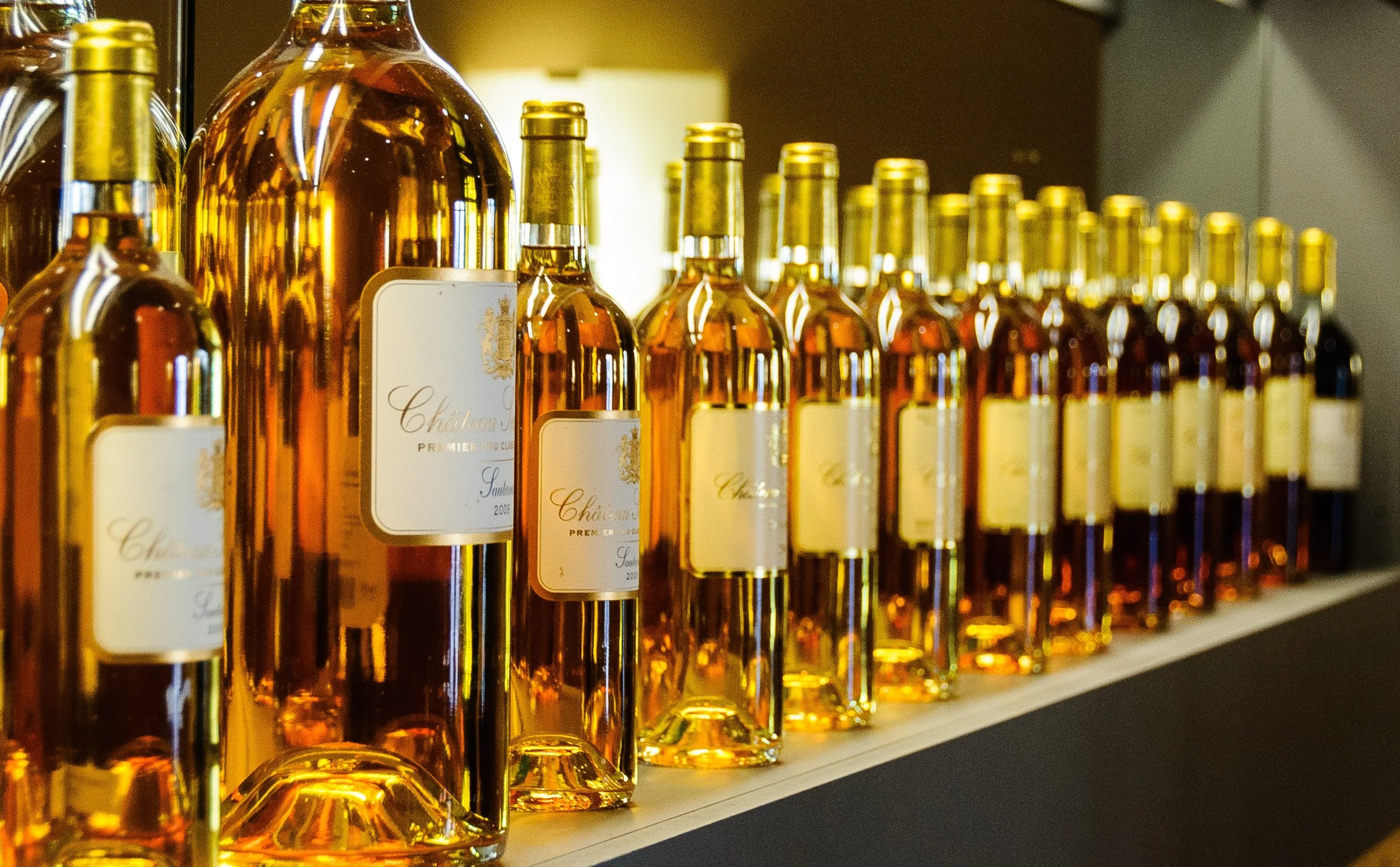A row of amber bottles on a shelf, with gold caps and white labels, reflecting light.