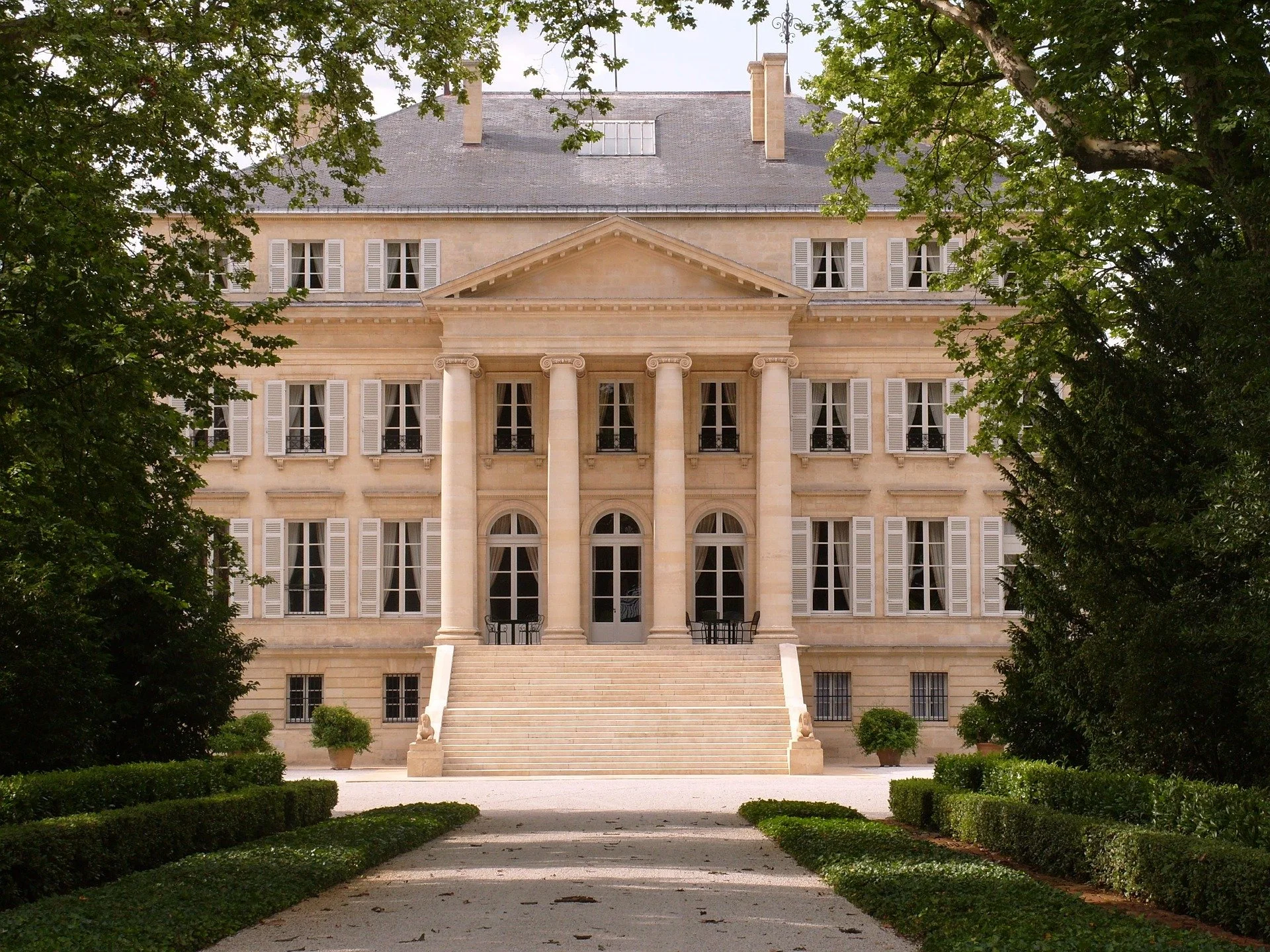 Front view of a large, elegant mansion with stone stairs leading up to a porch supported by four columns, surrounded by green trees and bushes.
