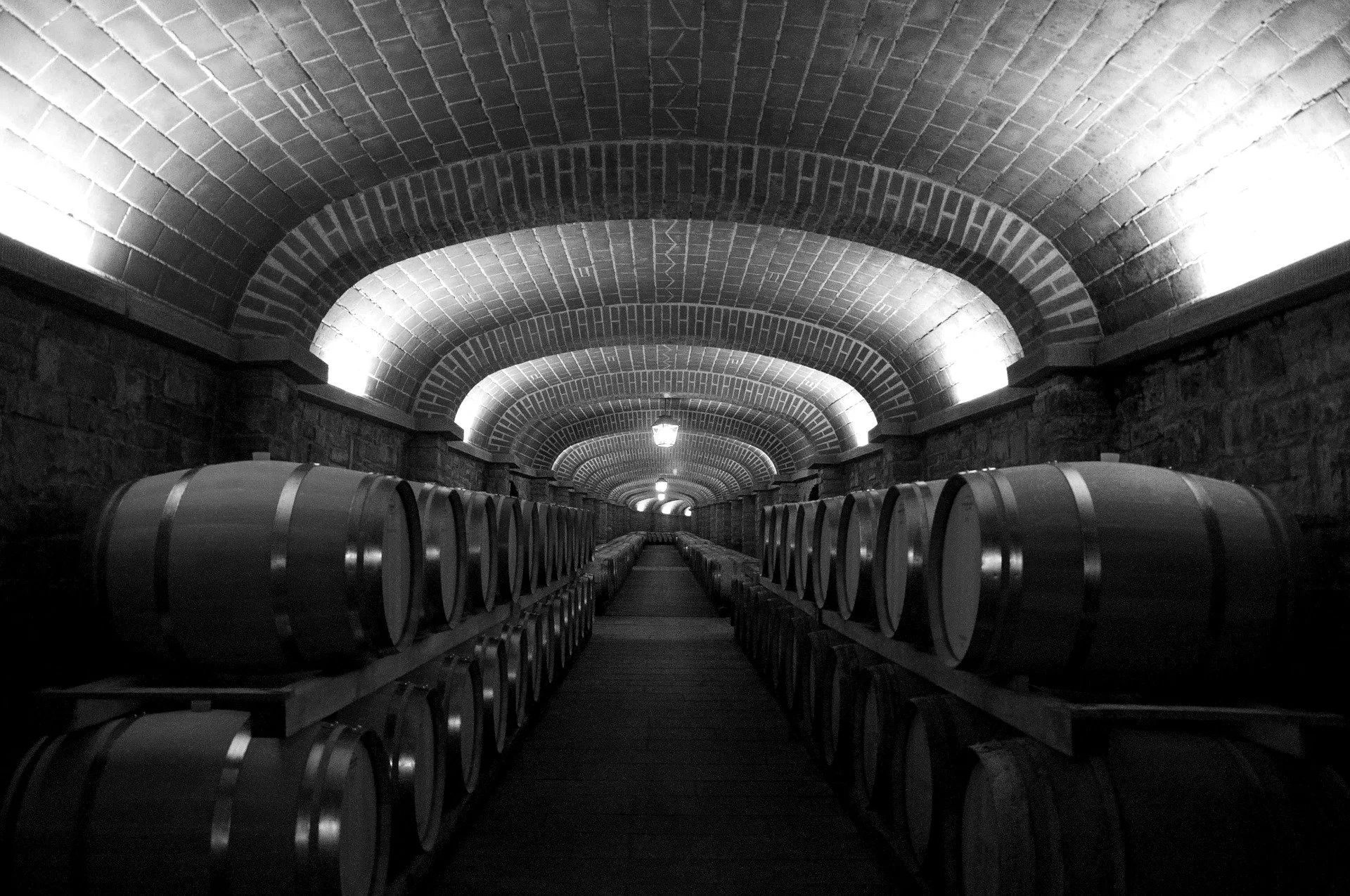 A tunnel made of brick arches with LED lighting, flanked by wine barrels on both sides, leading to a lit opening in the distance.