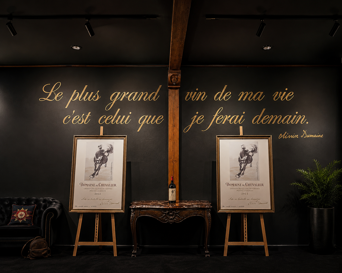 Interior wall with gold cursive French quote, two framed wine posters on easels, a small ornate table, a leather sofa with a crest pillow, a potted plant, and overhead track lighting.