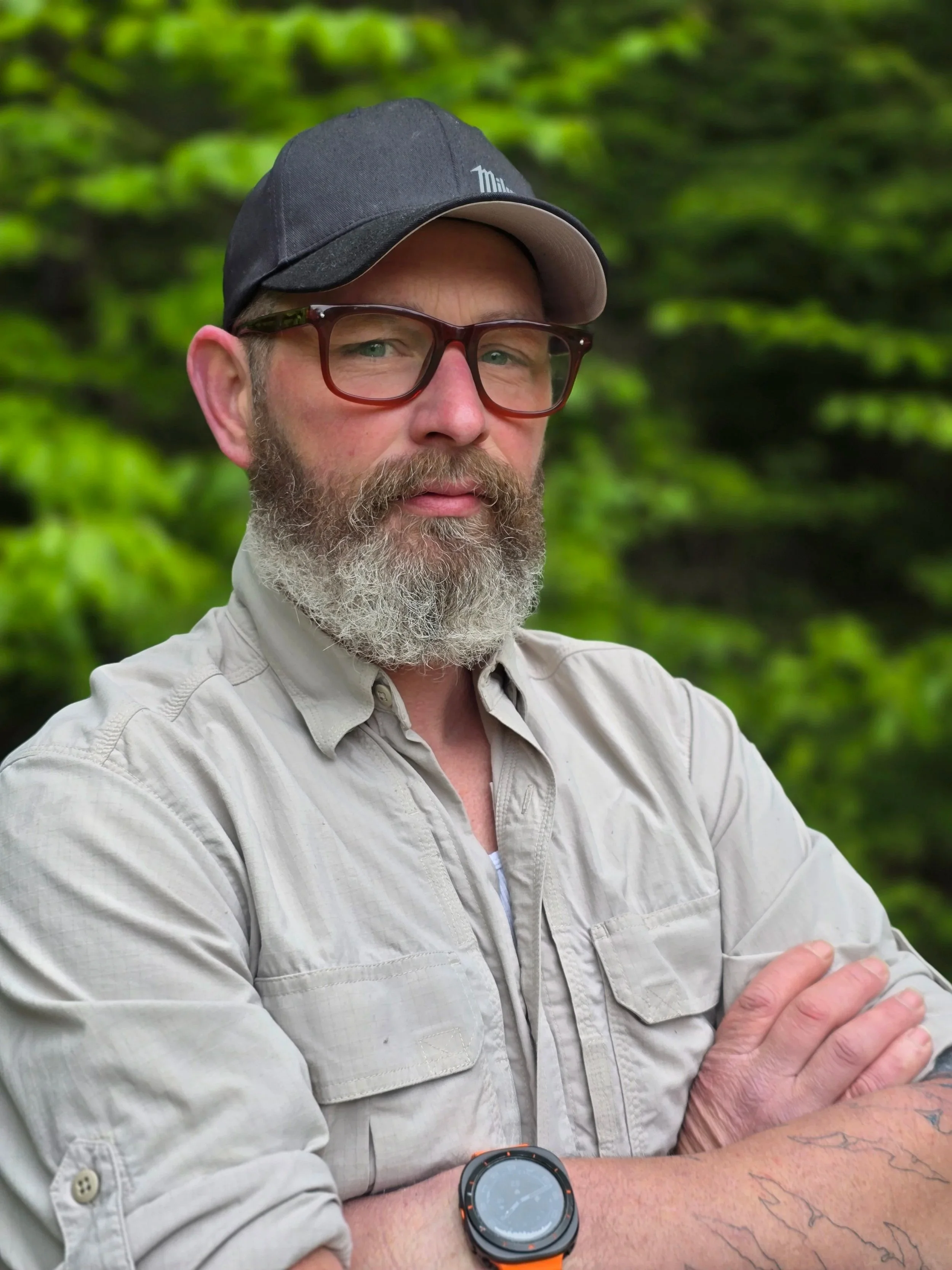 A man with glasses, a beard, and a mustache wearing a black cap and a beige shirt. He is standing outdoors with green foliage in the background, arms crossed, and wearing a black watch on his left wrist.