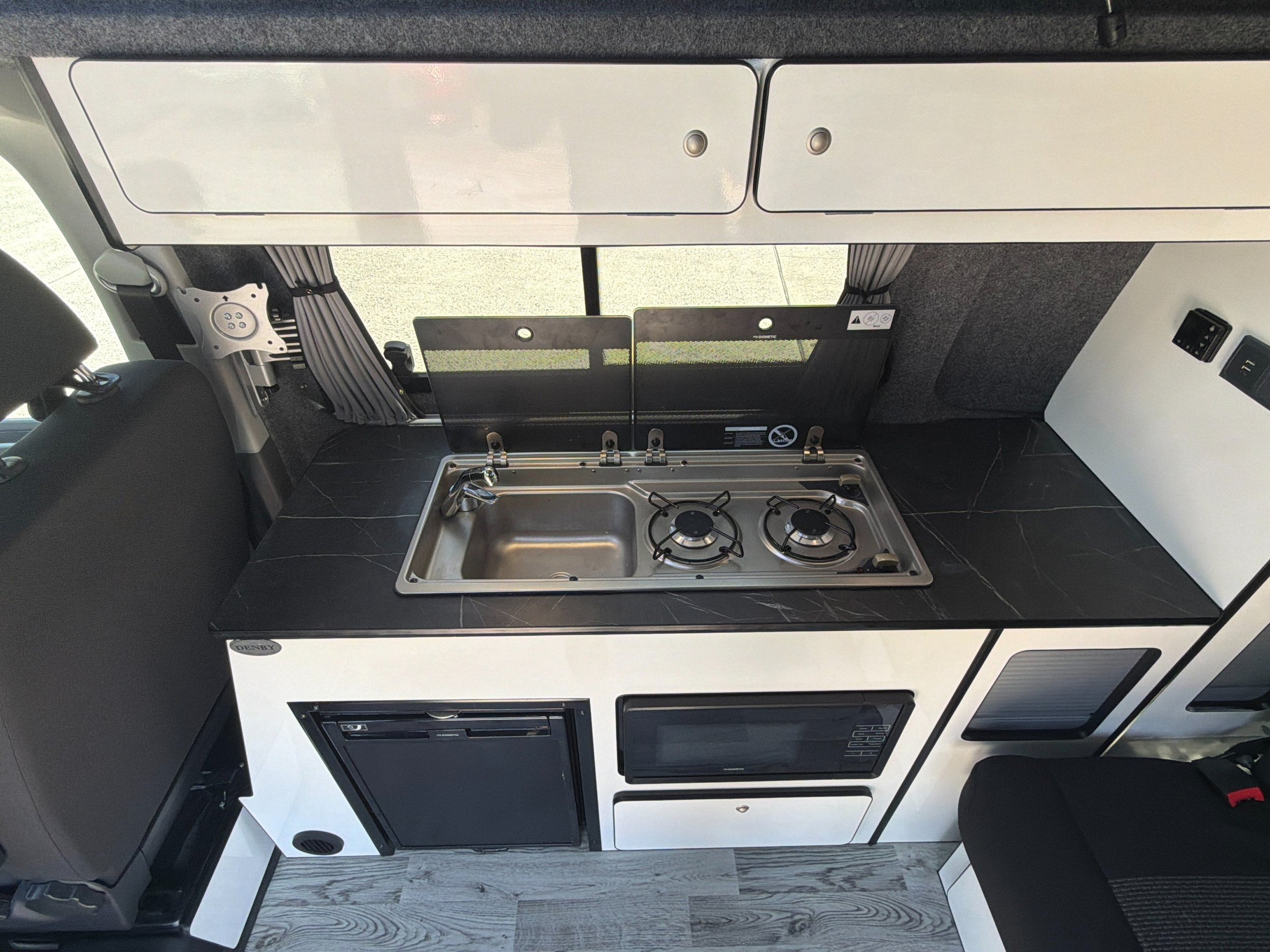 Fully equipped campervan kitchen area featuring a gas hob, sink, fridge, microwave, and storage cupboards.